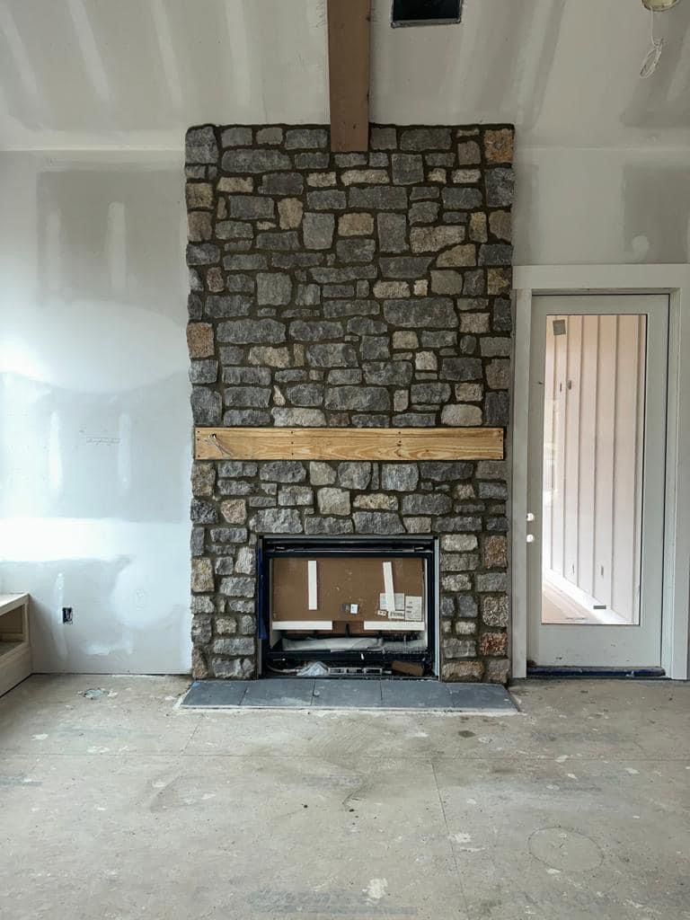 A rustic stone fireplace with a wooden mantel and beam under construction in a room with unfinished drywall and flooring.