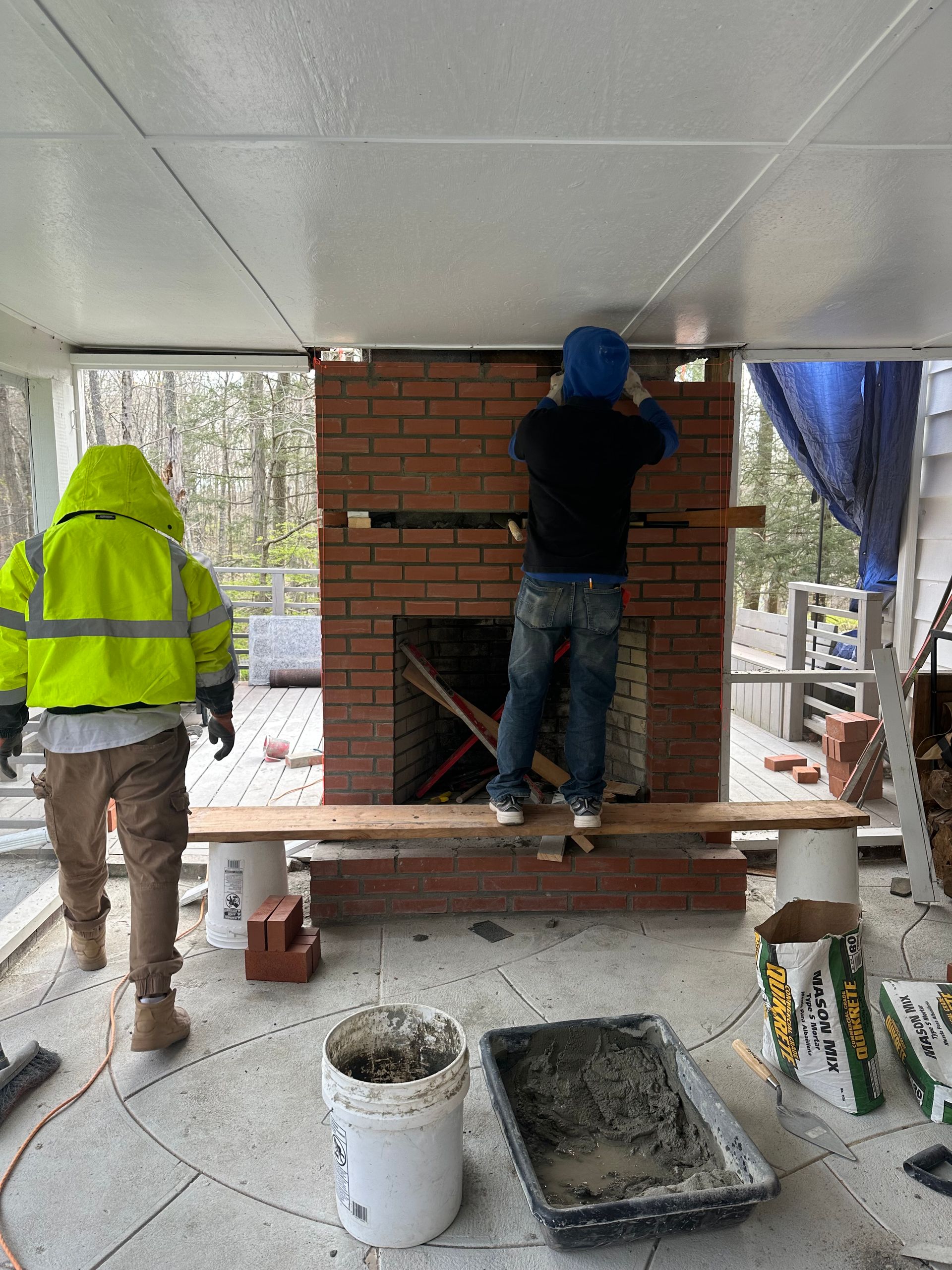 Two workers renovate an outdoor brick fireplace under a covered patio, with construction buckets and tools nearby.