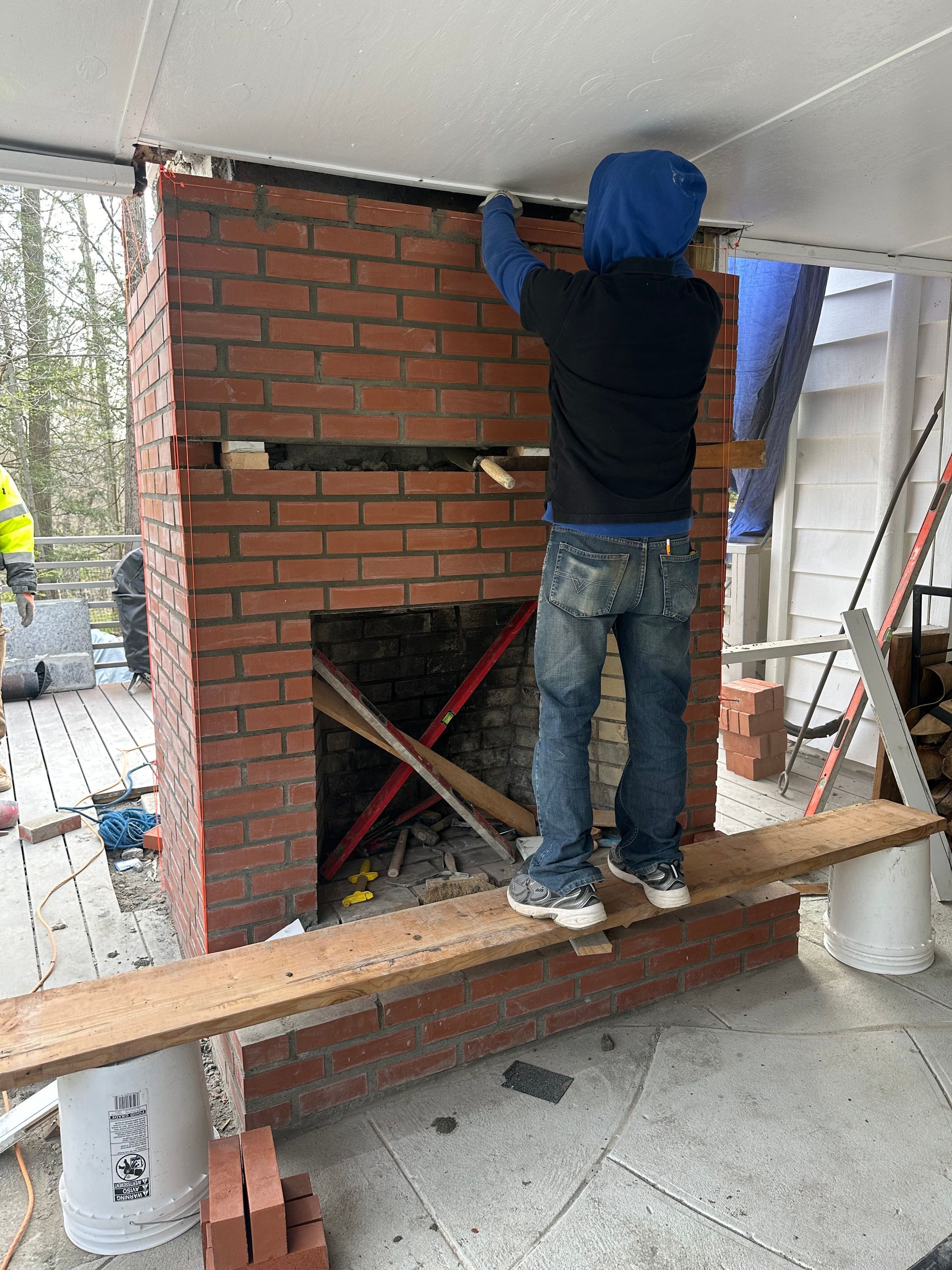A person in a blue hoodie stands on a wooden plank, working on a brick chimney construction under a porch roof.