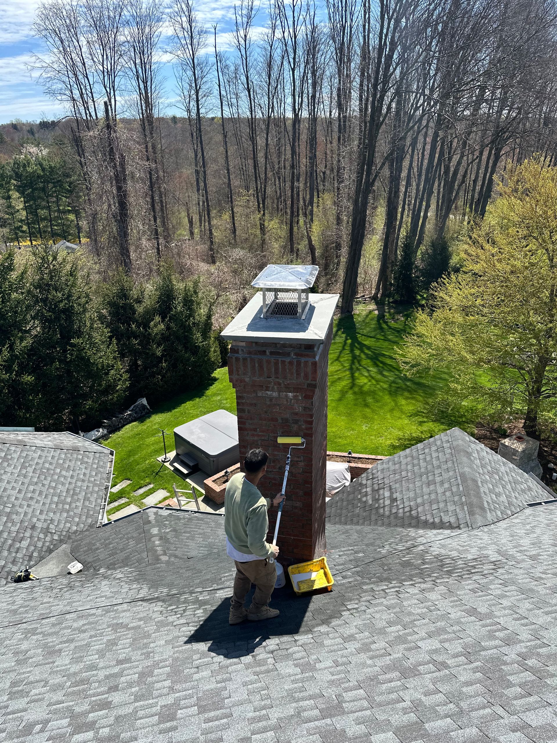 A person on a residential roof uses a roller to apply sealant to a tall brick chimney.