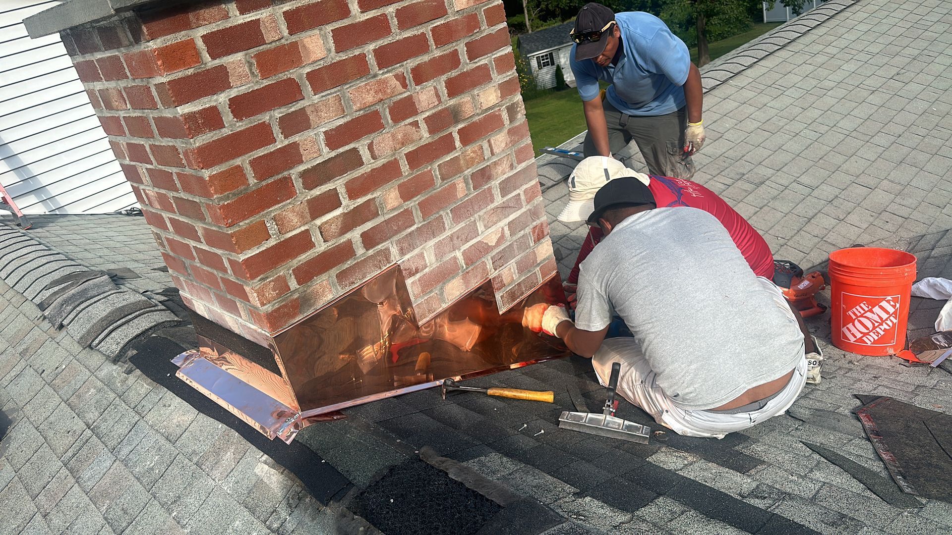 Two construction workers install copper flashing around a brick chimney on a shingled roof.
