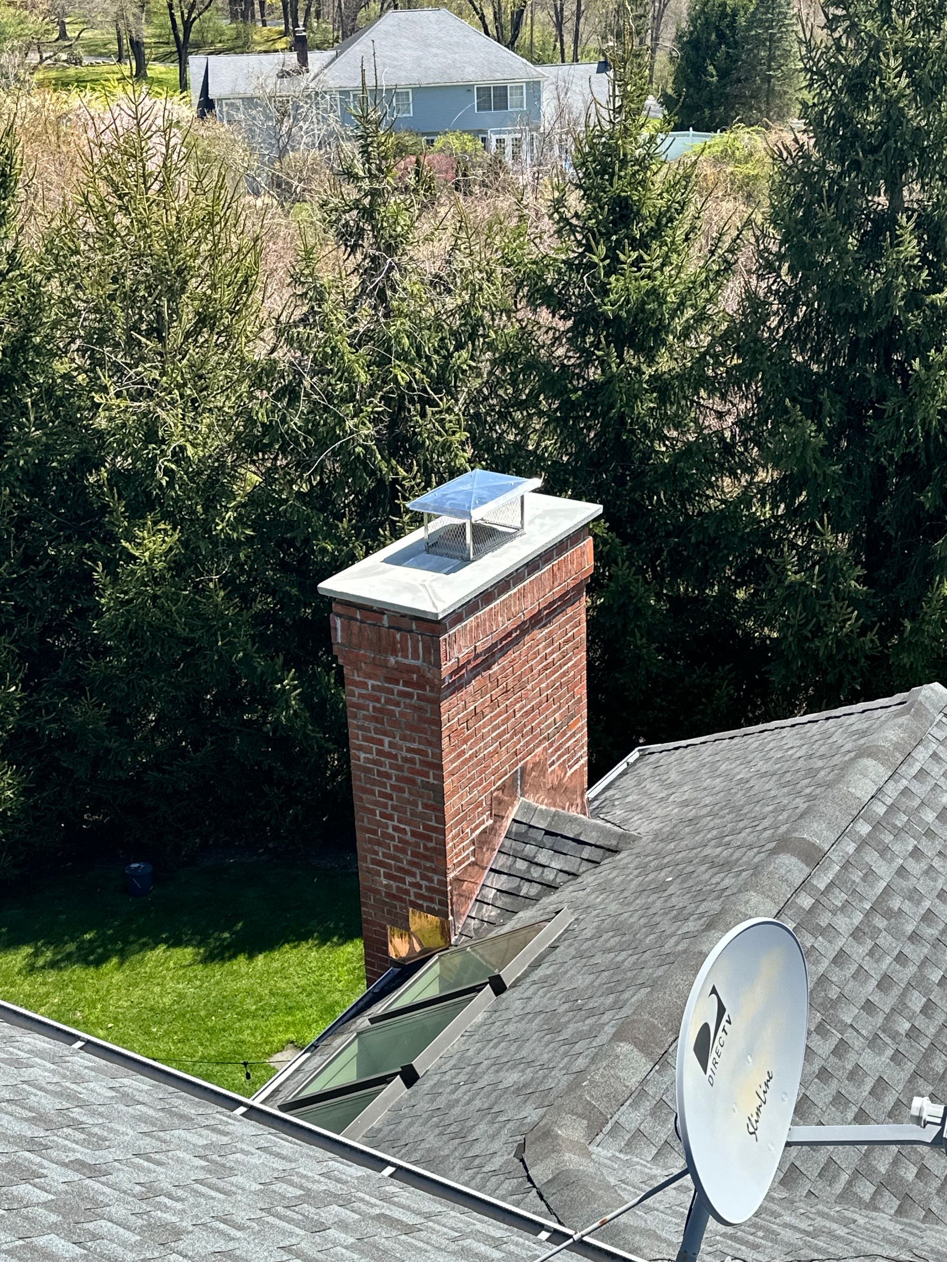 A brick chimney with a metal cap sits on a shingled roof next to a satellite dish, with trees and a house in the background.