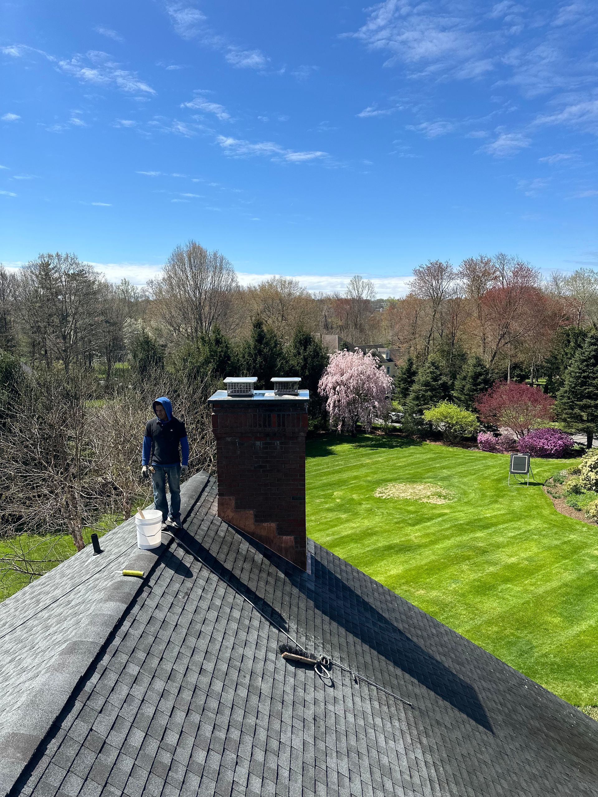A person standing on a sloped shingled roof next to a brick chimney overlooking a green lawn with blooming trees.