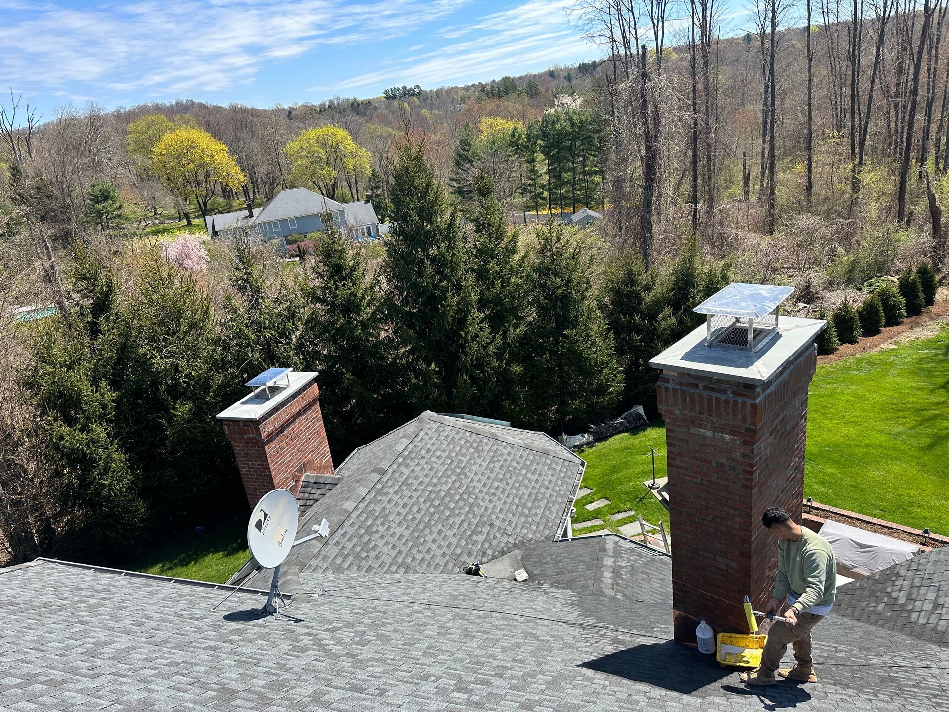 A person works on a grey shingled roof next to a brick chimney, with a satellite dish and scenic wooded yard nearby.