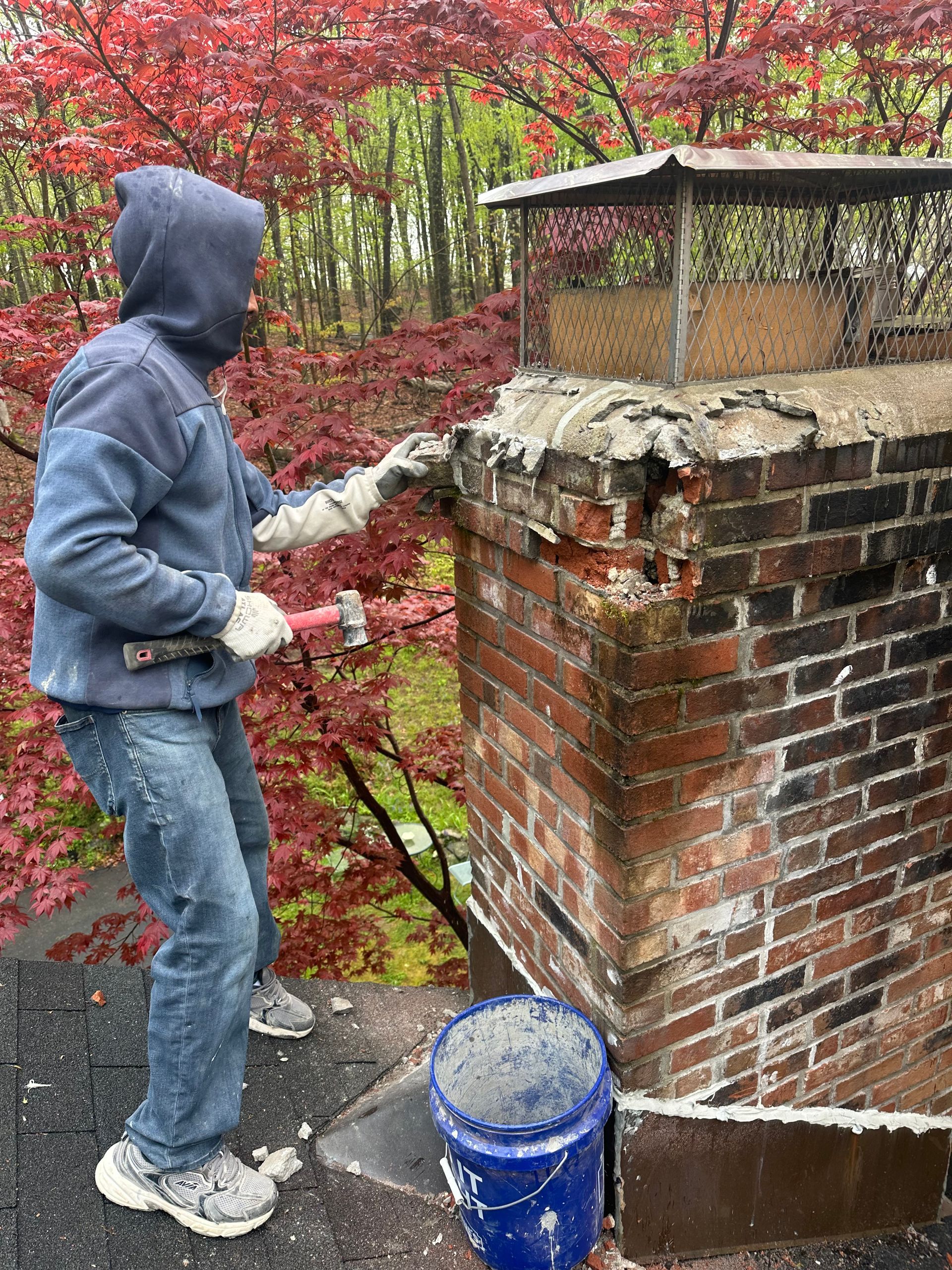 A worker in a blue hoodie and jeans uses a hammer to repair a crumbling brick chimney on a rooftop.