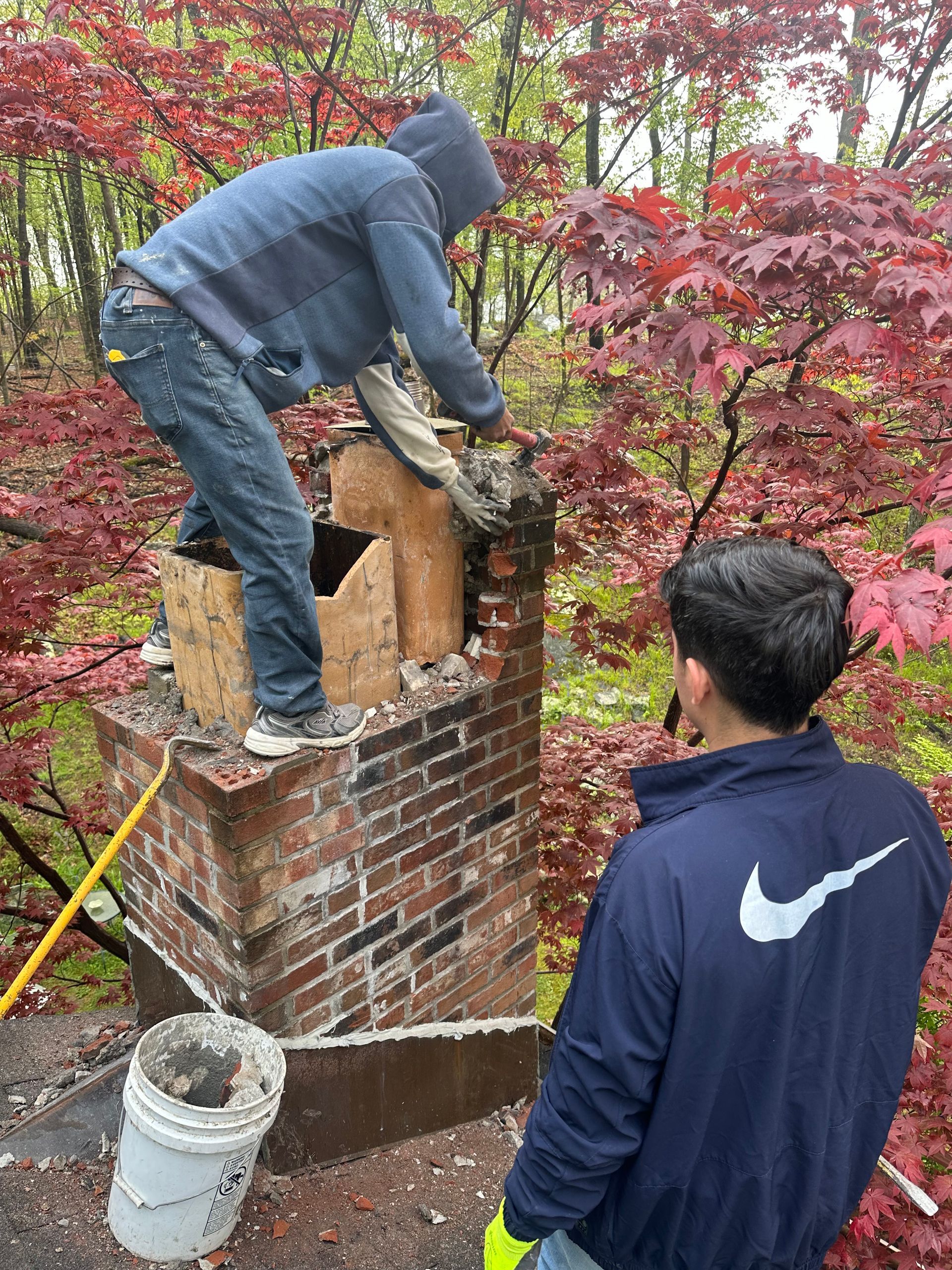 Two people perform maintenance on a brick chimney outdoors surrounded by trees with autumn foliage.