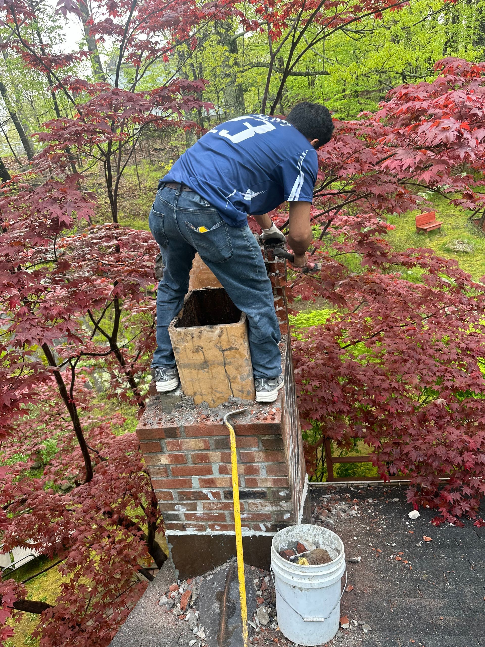 A worker standing on a chimney roof, repairing the brick structure while surrounded by red-leafed trees.
