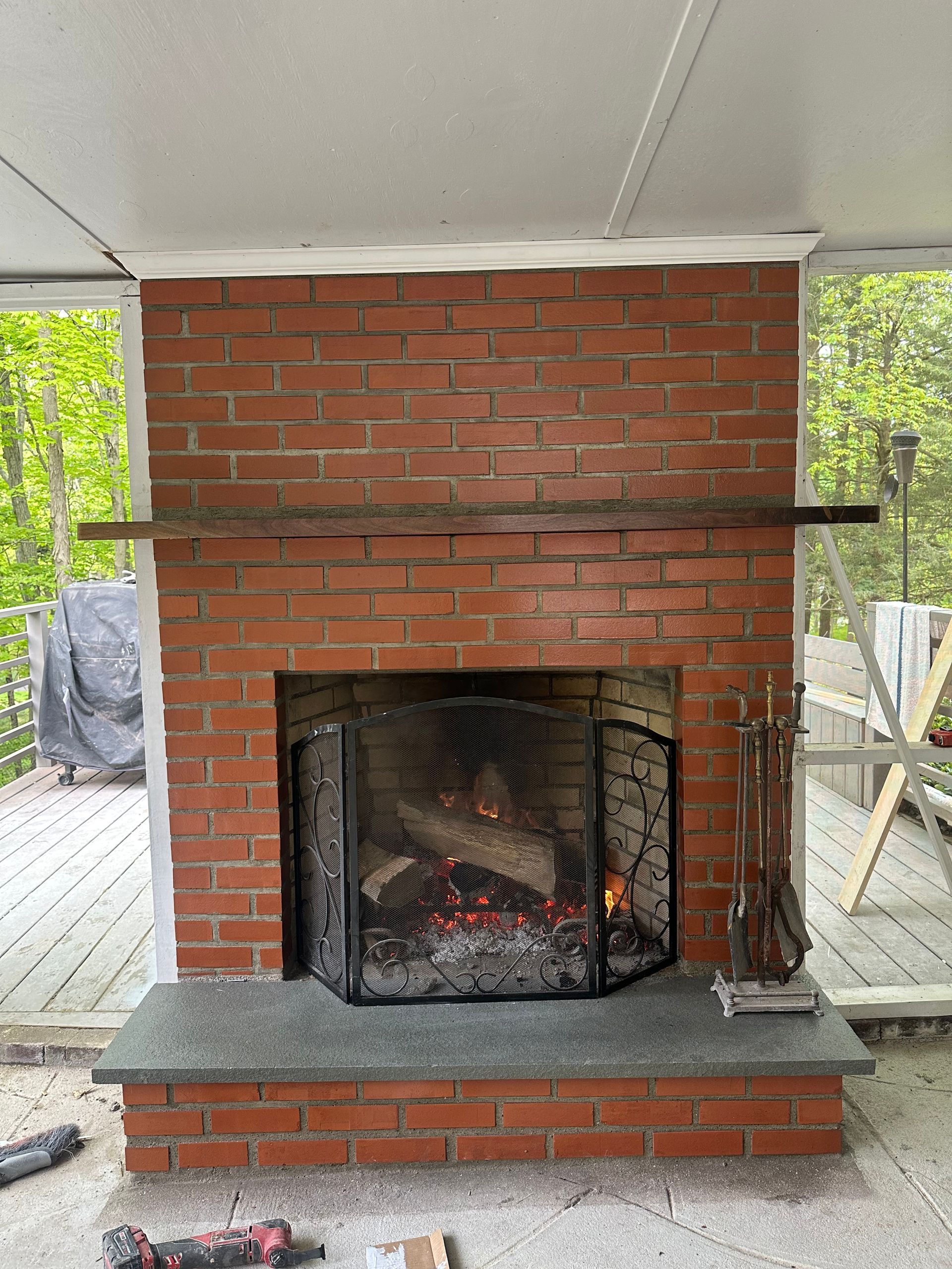 A red brick fireplace with a stone hearth, a fireplace screen, and a rustic wooden mantel on an outdoor porch.