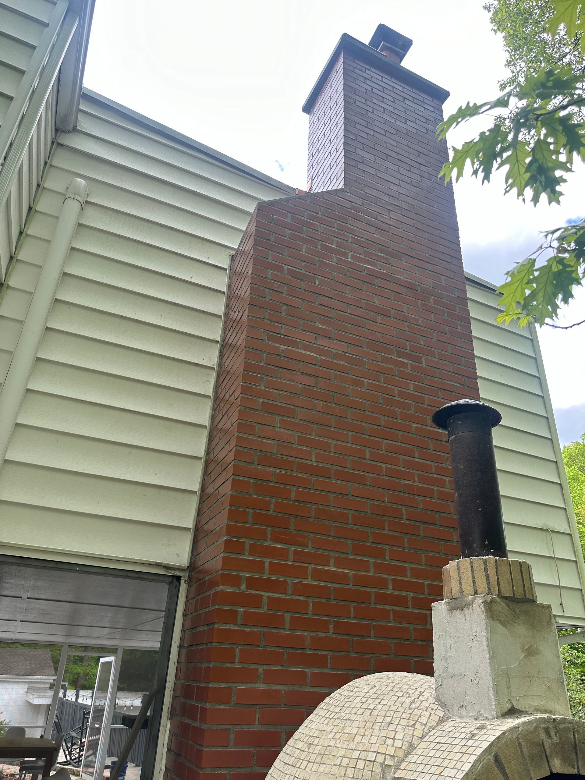 A tall brick chimney stands against light-colored house siding, with a stone outdoor pizza oven in the foreground.