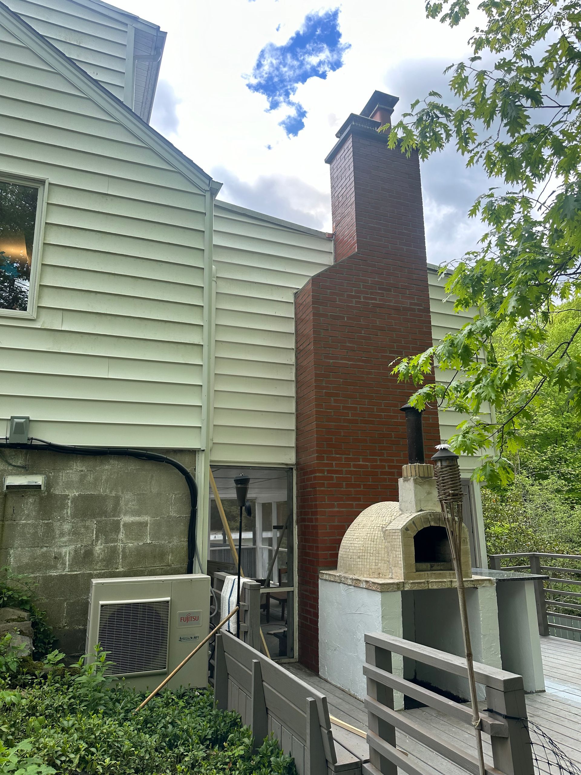 A brick chimney and outdoor stone pizza oven on a deck next to a light-sided house with green foliage.
