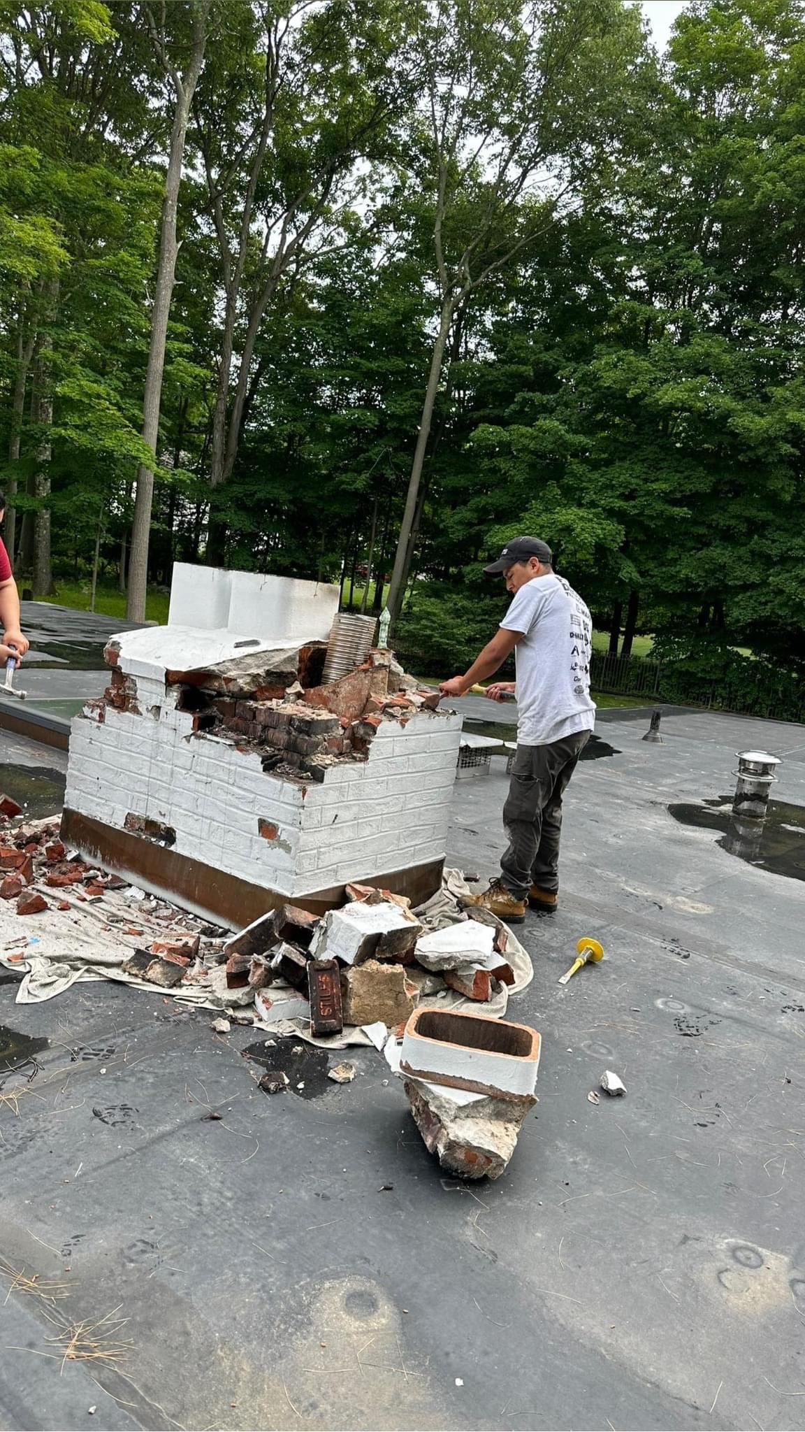 A worker in a white t-shirt uses a tool to demolish a brick chimney structure on a flat, gravel-covered roof outdoors.
