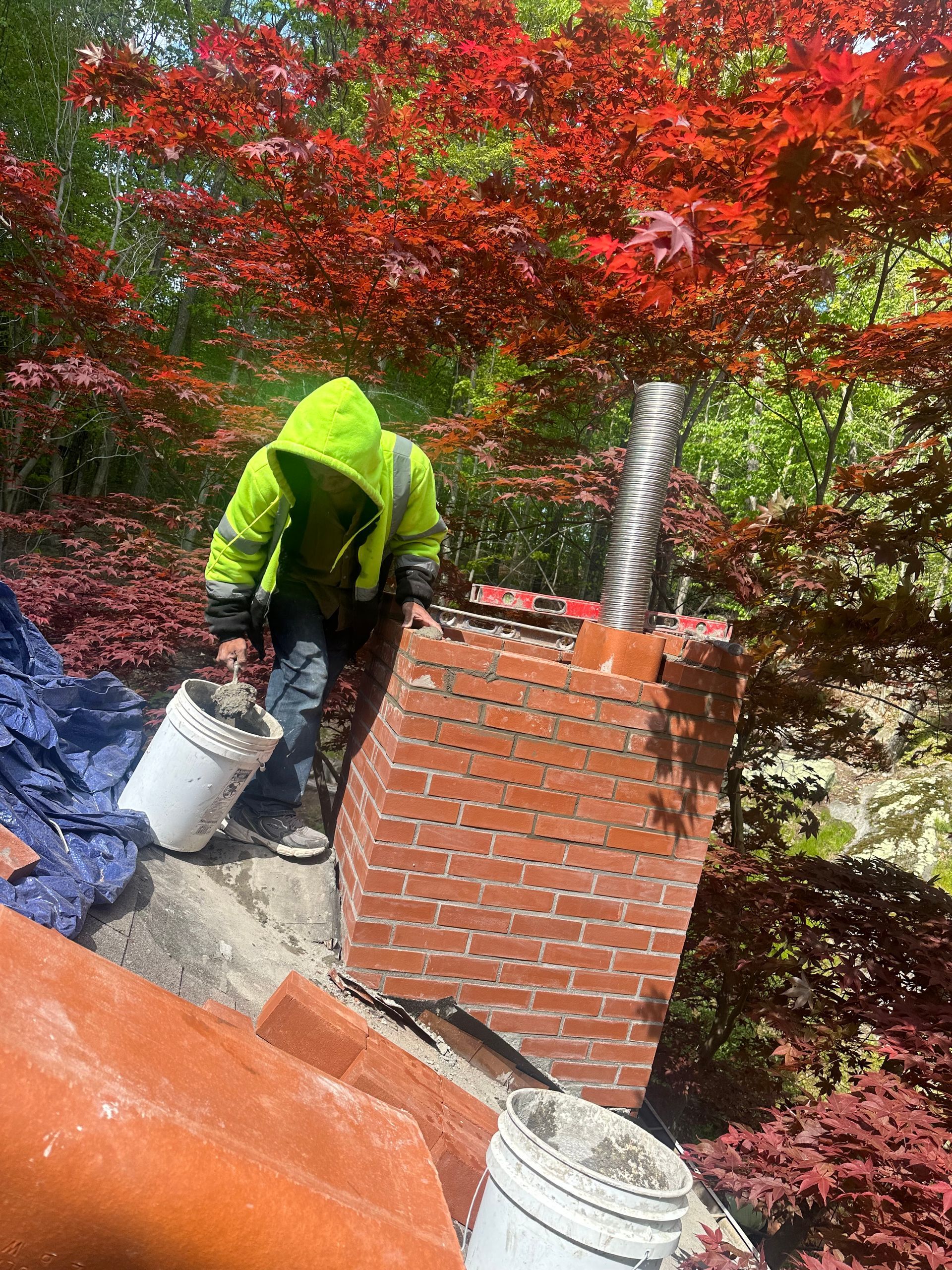 A construction worker in a high-visibility lime jacket works on a brick chimney with a metal flue on a rooftop.