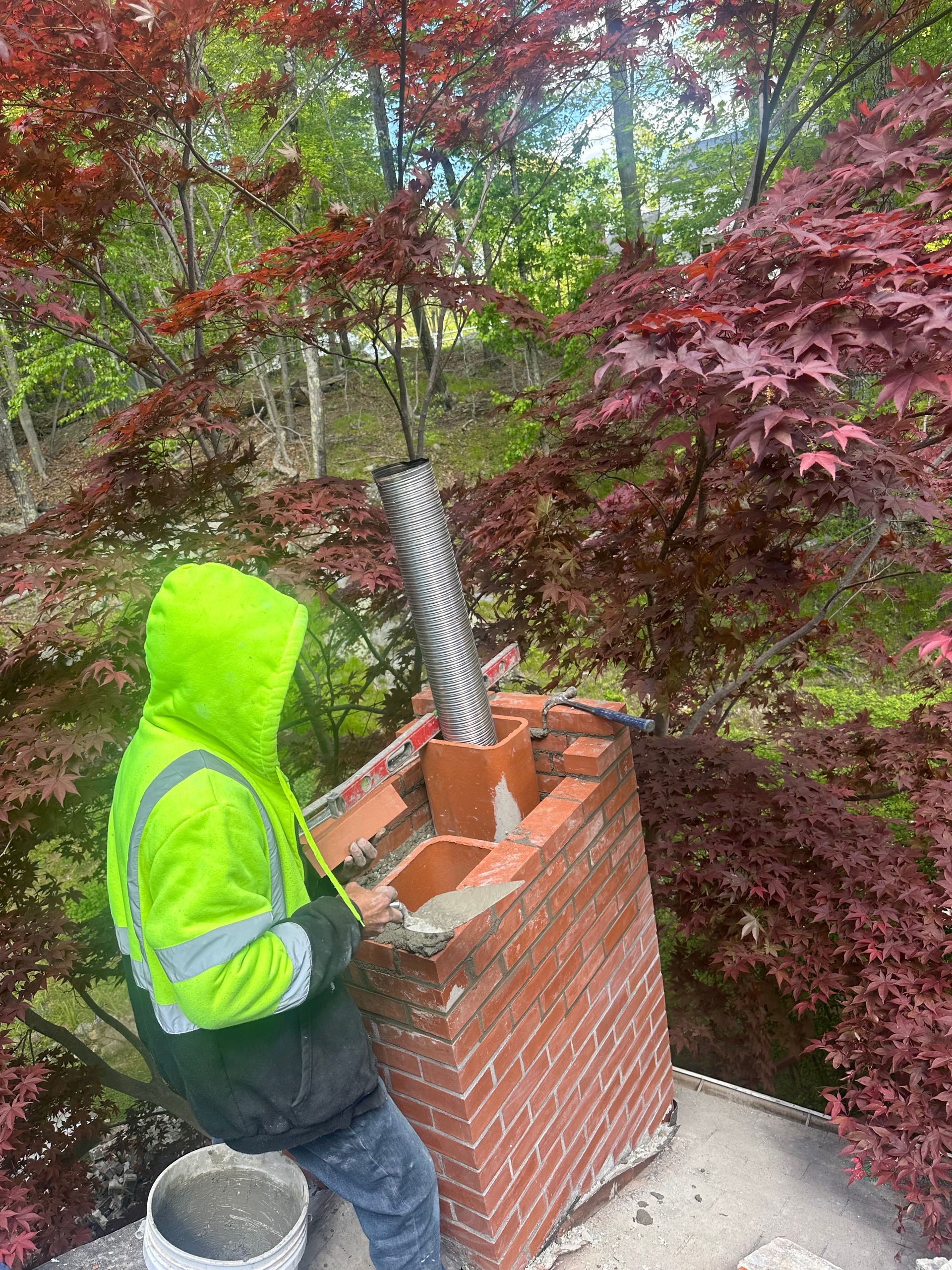 A worker in a high-visibility jacket installs a metal chimney liner into a brick chimney outdoors among red-leaf trees.