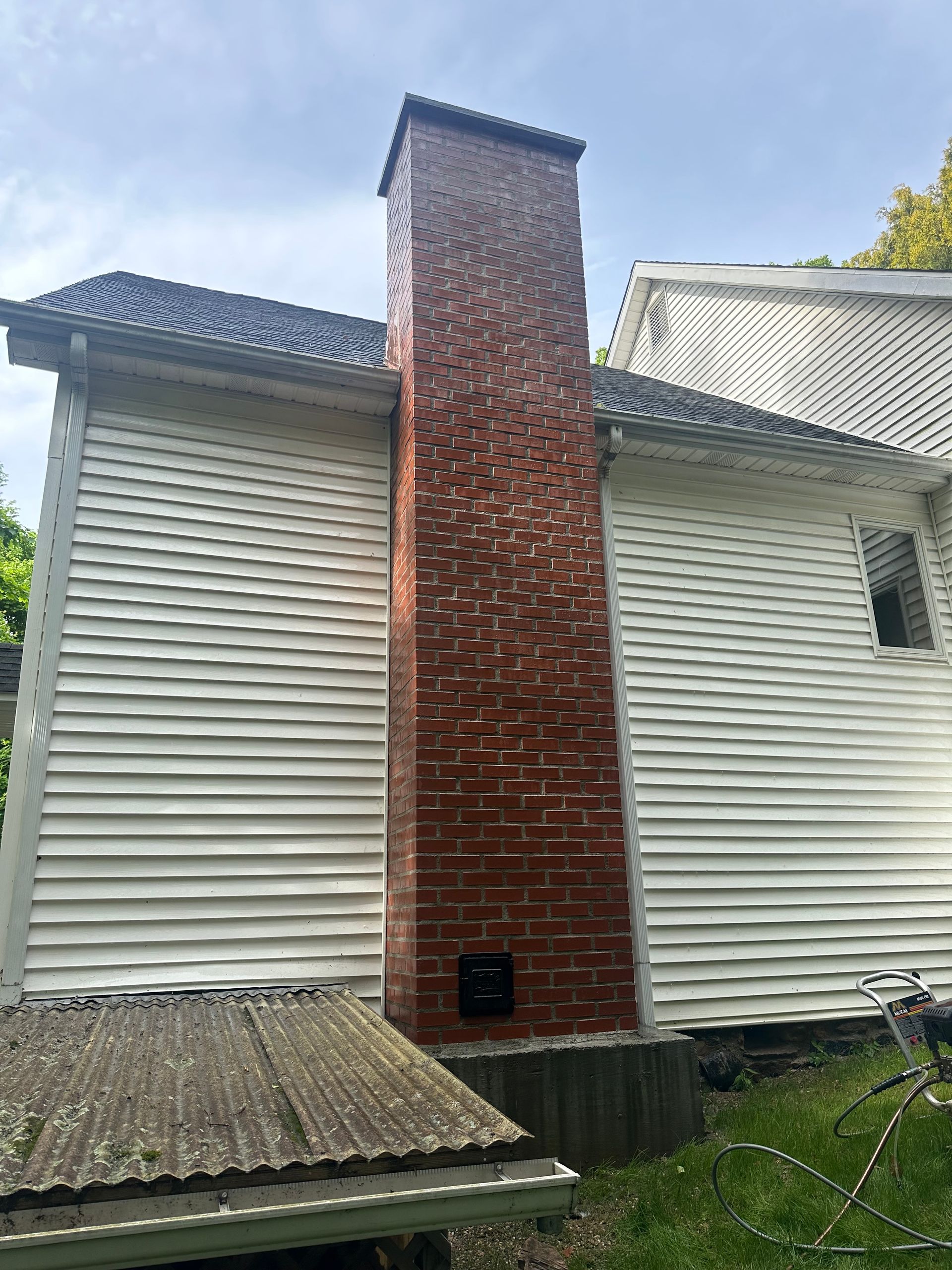 A red brick chimney rises from the ground against the white vinyl siding of a two-story house exterior.