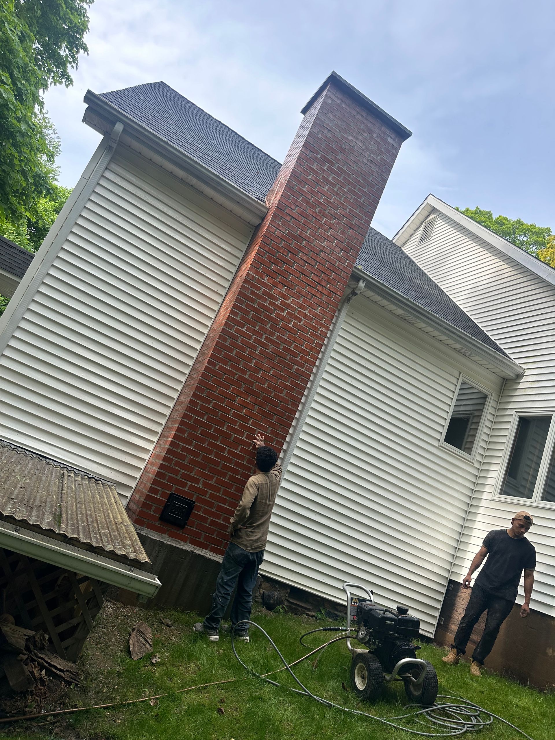 Two people power wash a large brick chimney on the side of a house with white siding.