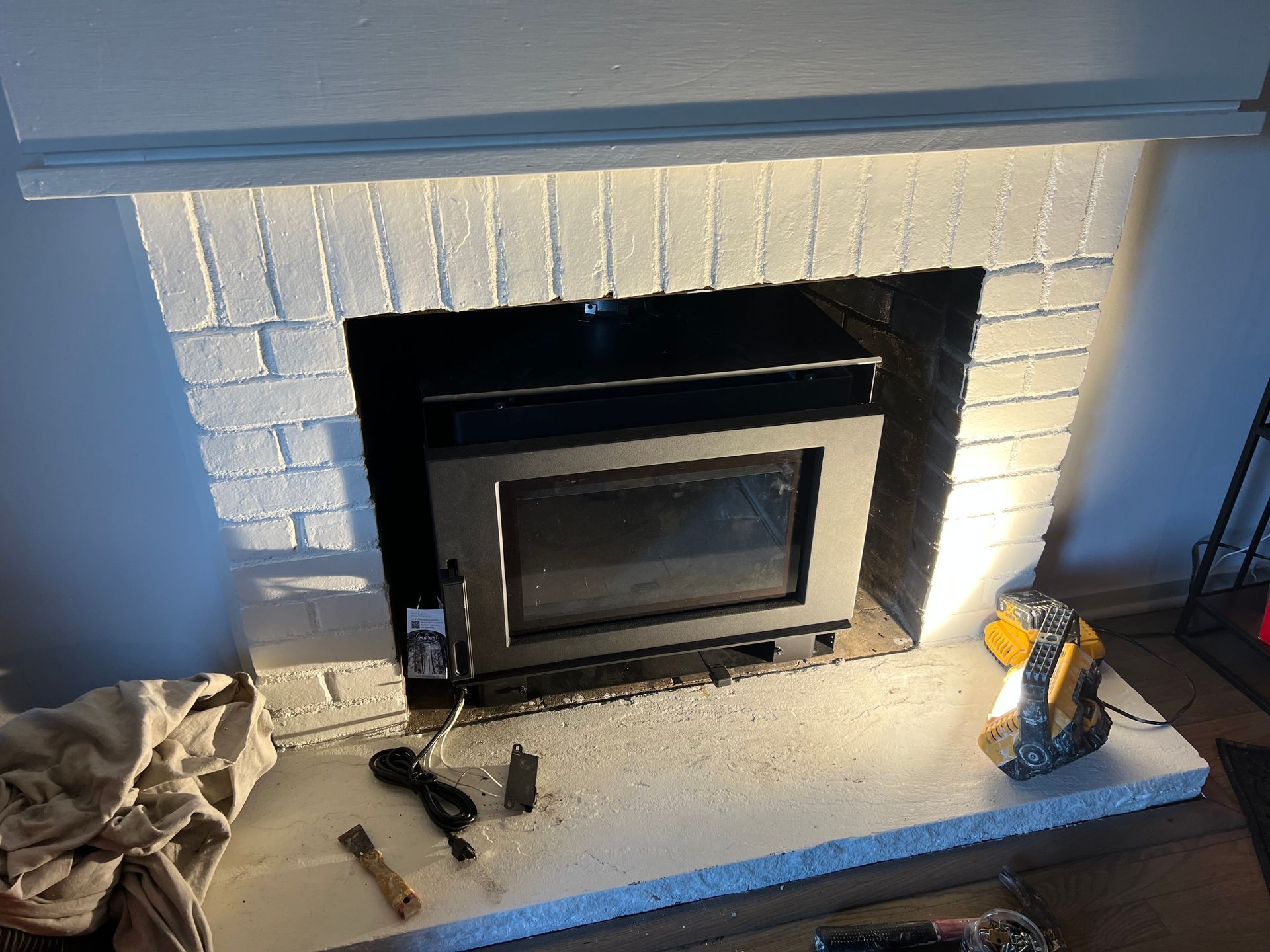 A fireplace insert installed in a white-painted brick fireplace, with a small brush and power cord on the hearth.