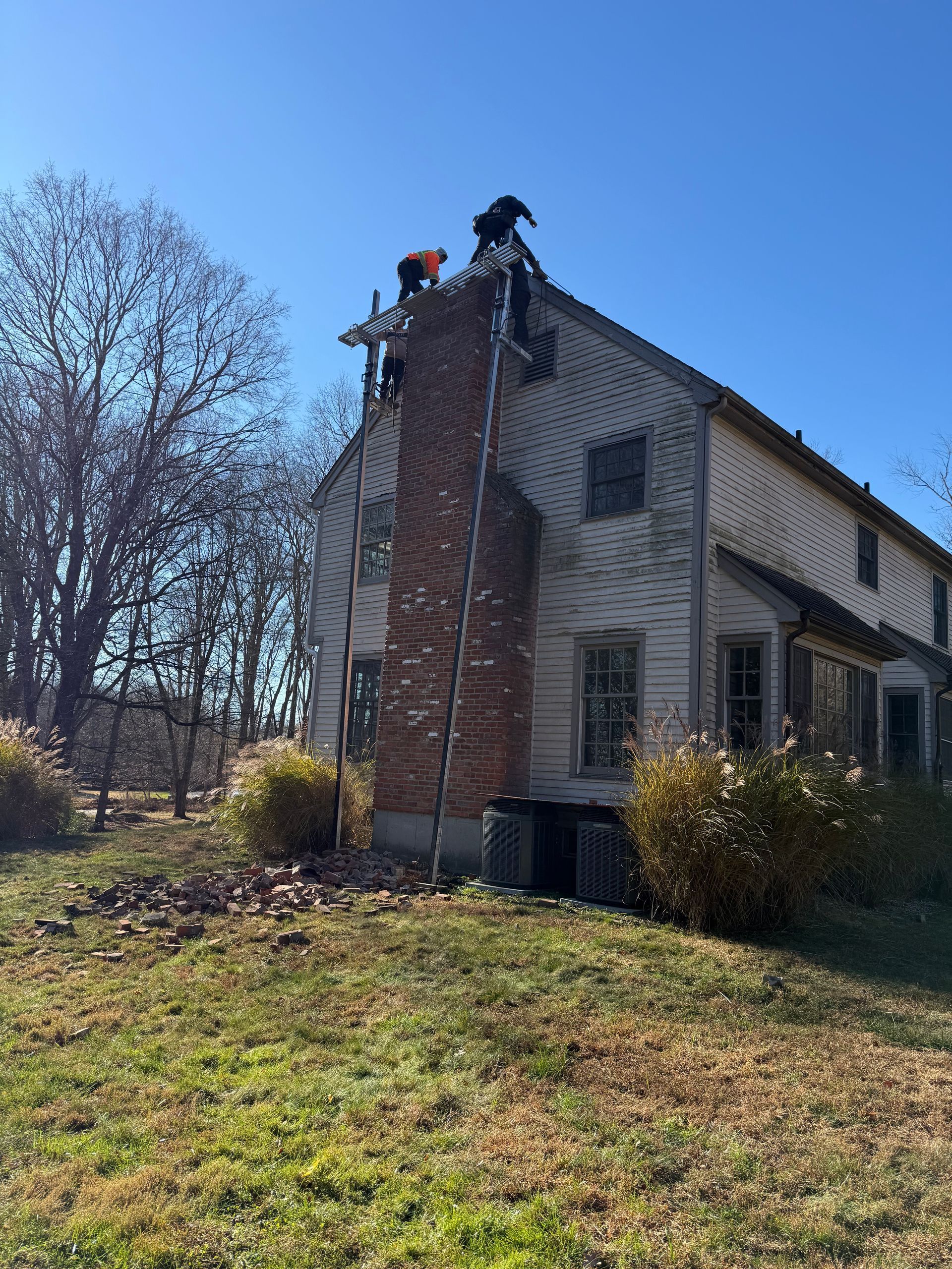 Two people on ladders work to dismantle the brick chimney on the side of a two-story house under a clear blue sky.