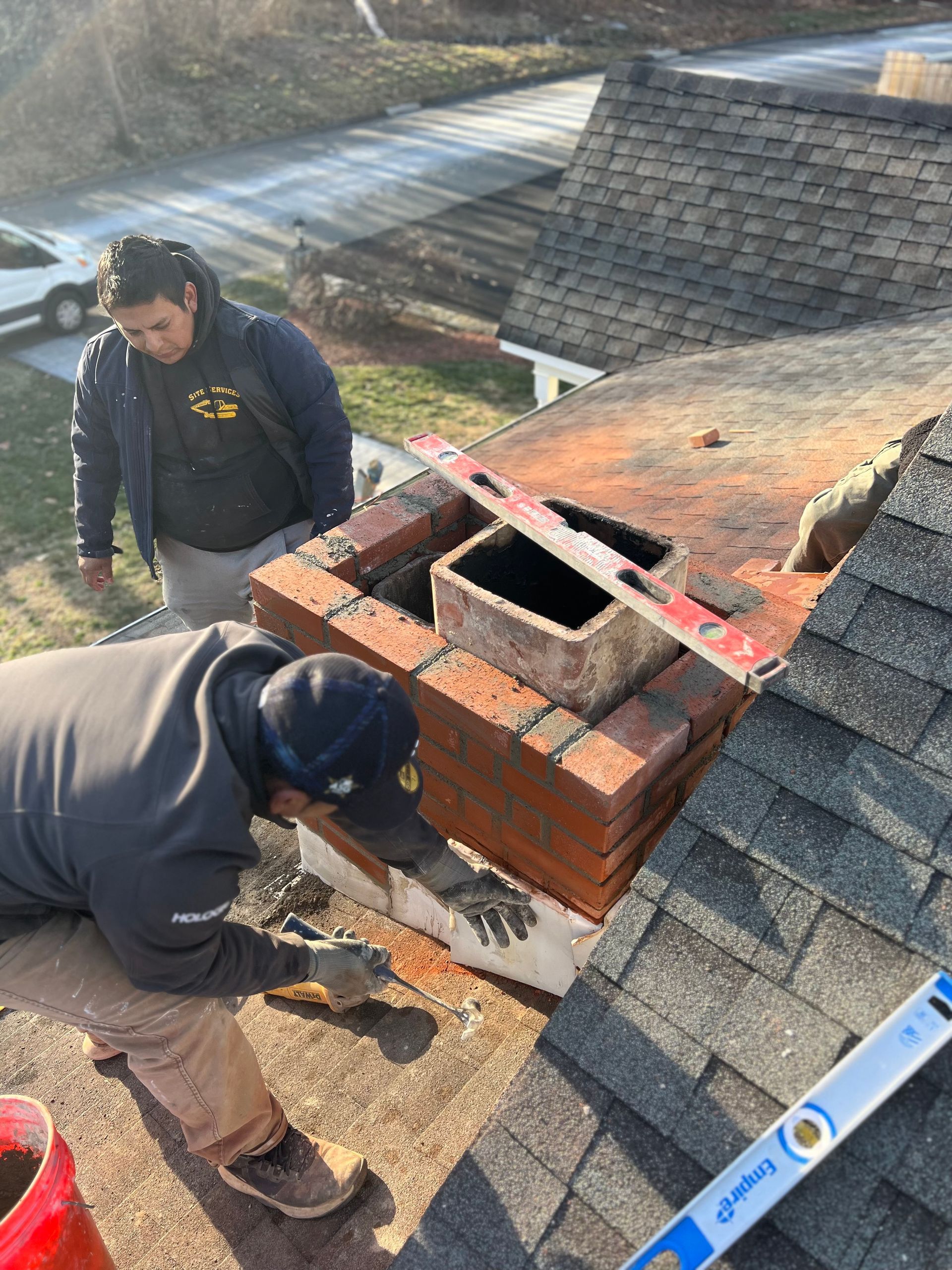 Two workers repairing a brick chimney on a residential roof. One worker applies mortar while the other observes.