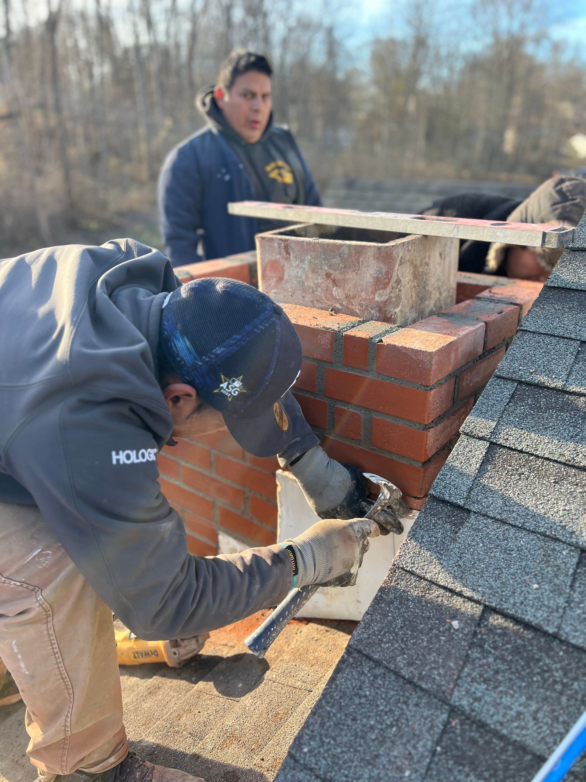 Two workers repair a brick chimney on a shingled roof, with one focused on the masonry and the other observing nearby.