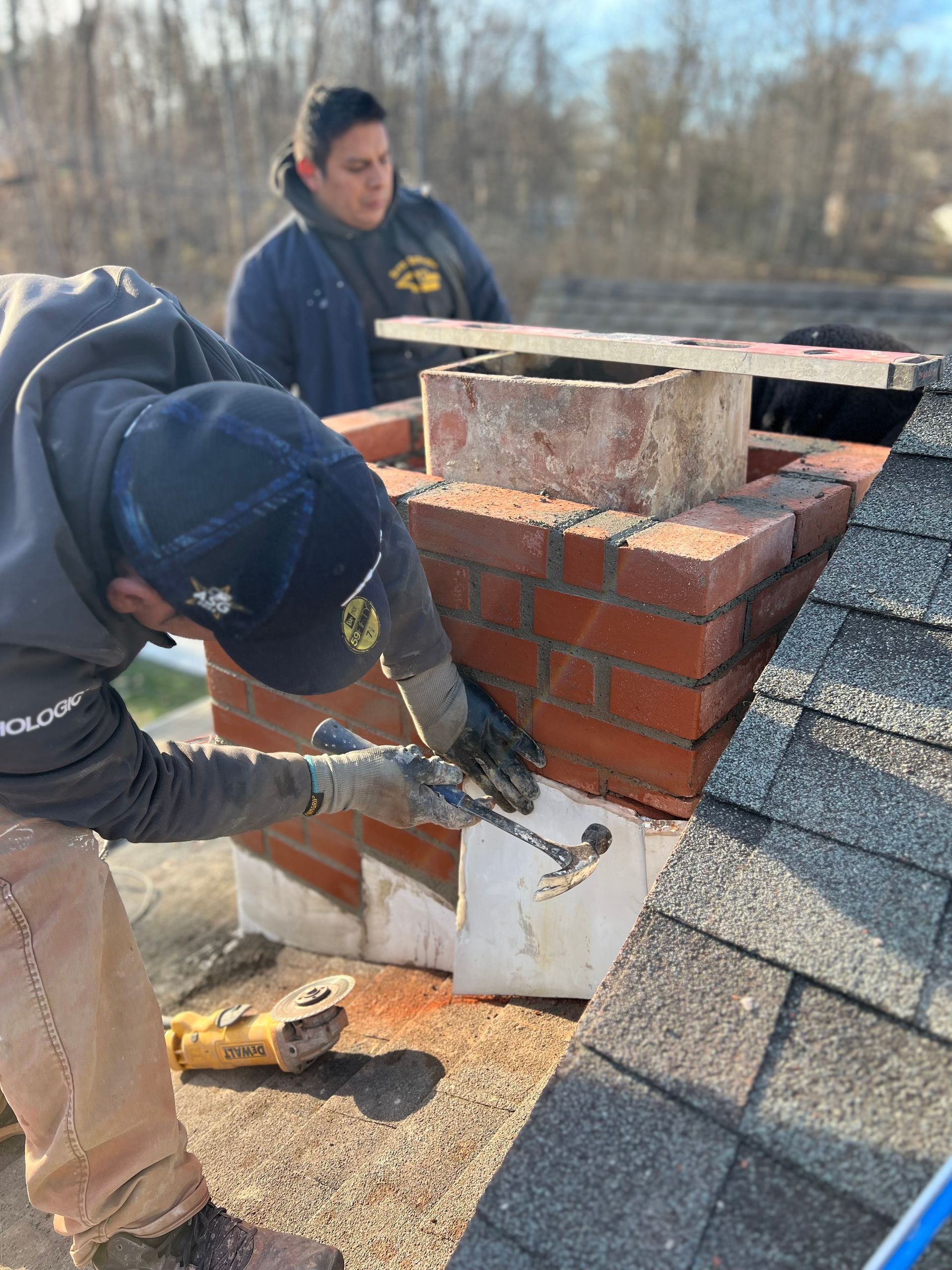 Two workers repair a brick chimney on a roof, one using a hammer on white flashing while the other looks on.