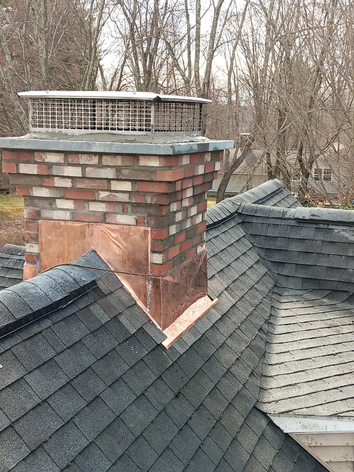 A brick chimney with a metal cap and copper flashing stands on a dark shingled roof surrounded by bare trees.