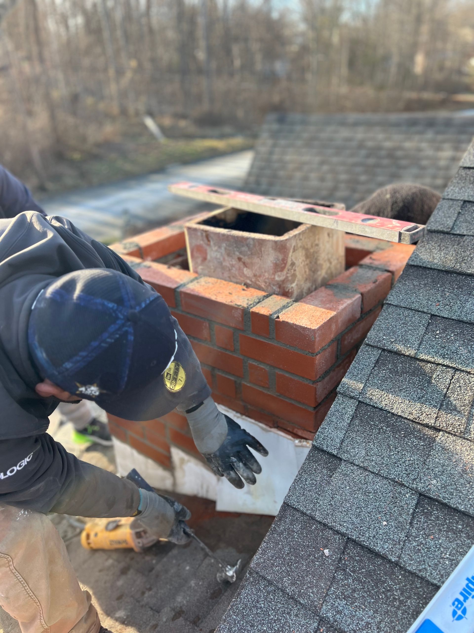 A person in work gear repairs a red brick chimney on a shingled residential roof.
