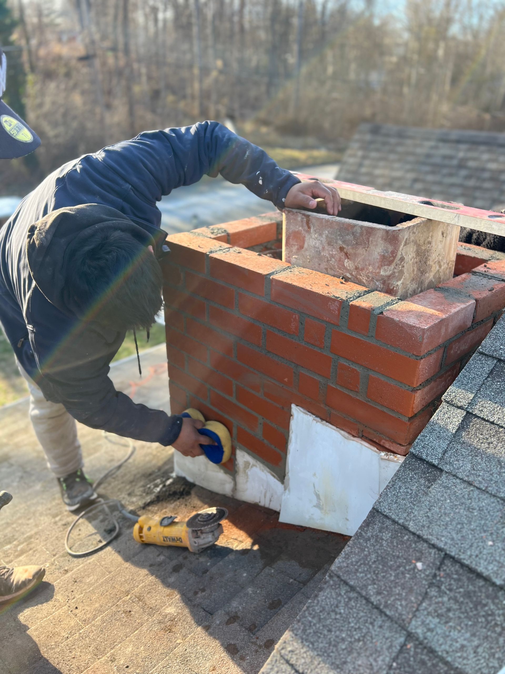 A person on a roof uses a power tool to smooth or clean the brickwork of a chimney stack.
