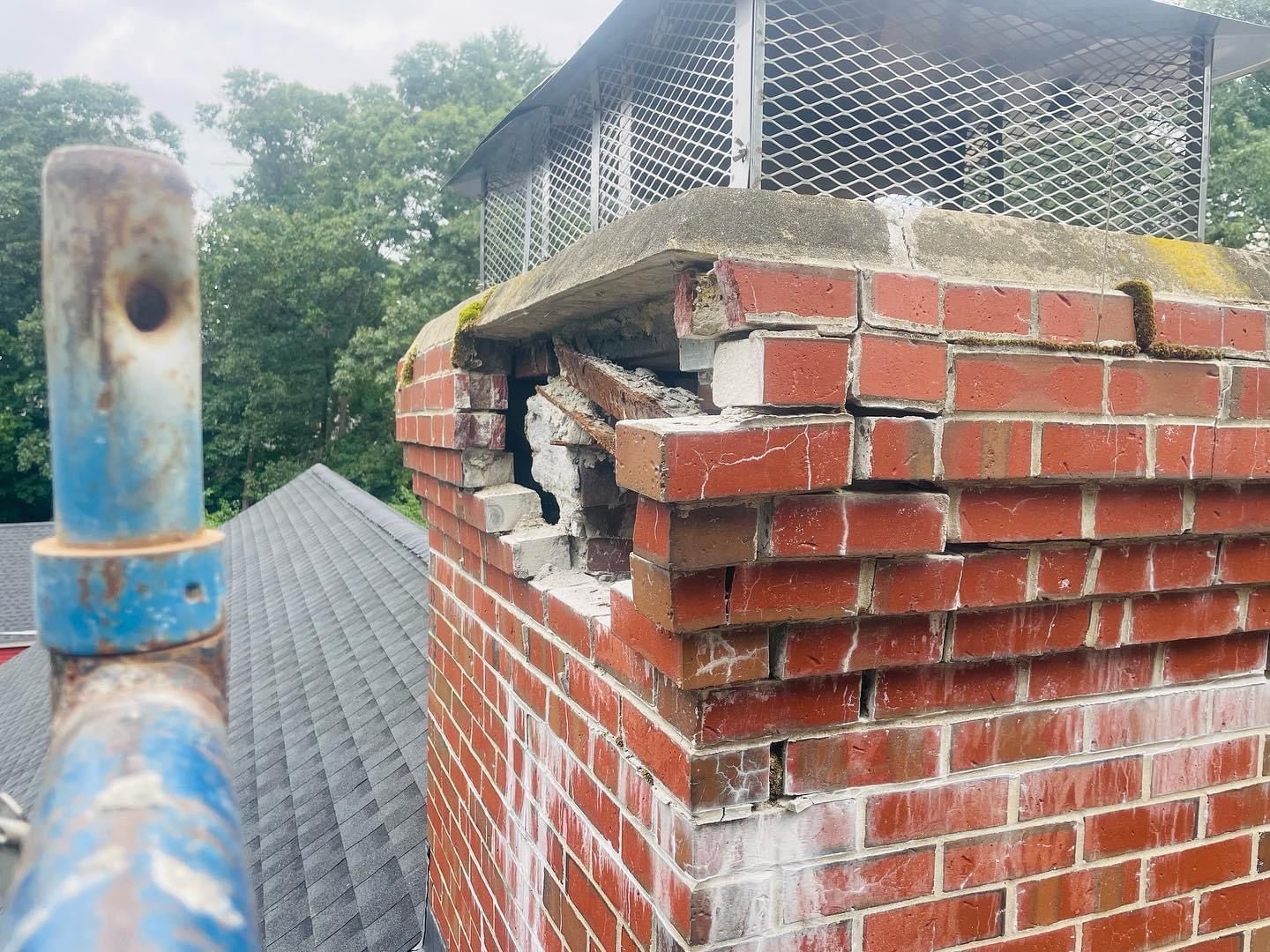 A damaged red brick chimney with a hole near the top, viewed from a rooftop next to a blue metal scaffolding pole.