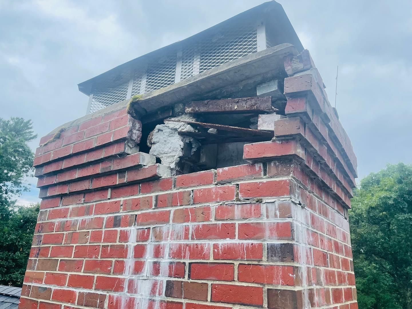 A damaged brick chimney with a large section missing near the top, exposing the inner structure and metal support beams.