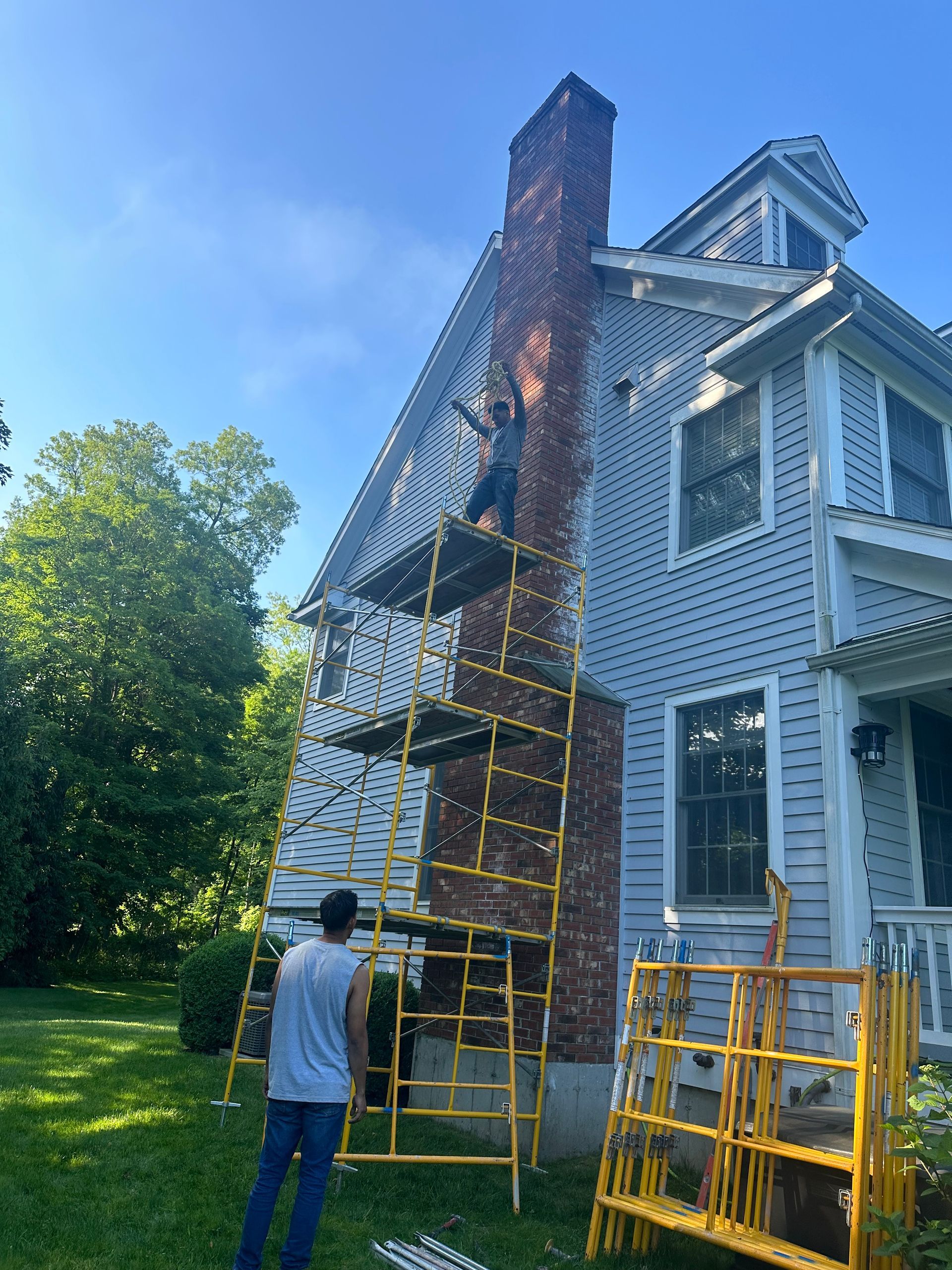 Workers use yellow scaffolding to repair a brick chimney on the side of a gray house in a grassy yard under a blue sky.