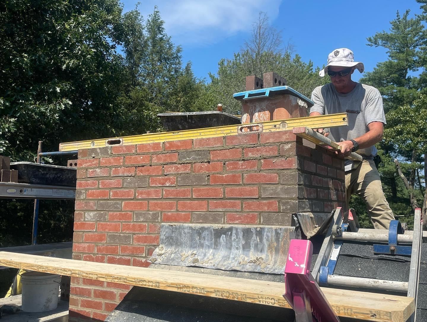A worker wearing a sun hat uses a level to measure the top of a brick chimney on a rooftop.