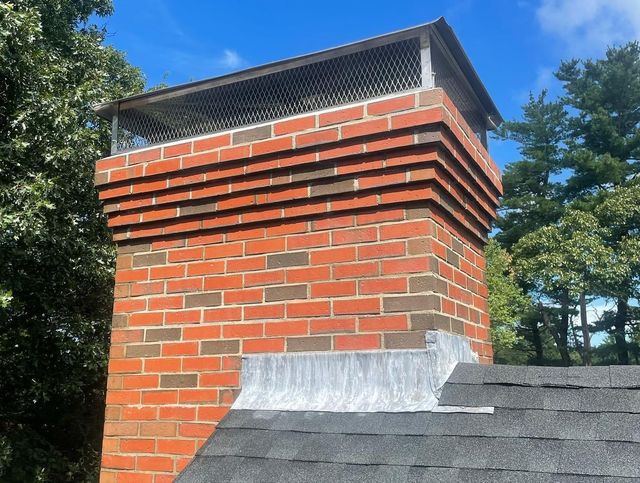 A brick chimney on a shingled roof with metal flashing at the base and a protective mesh cap at the top.