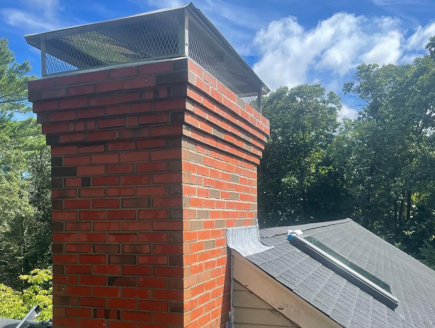 A red brick chimney with a metal mesh cap, situated next to a shingled roof with a vent on a sunny day.