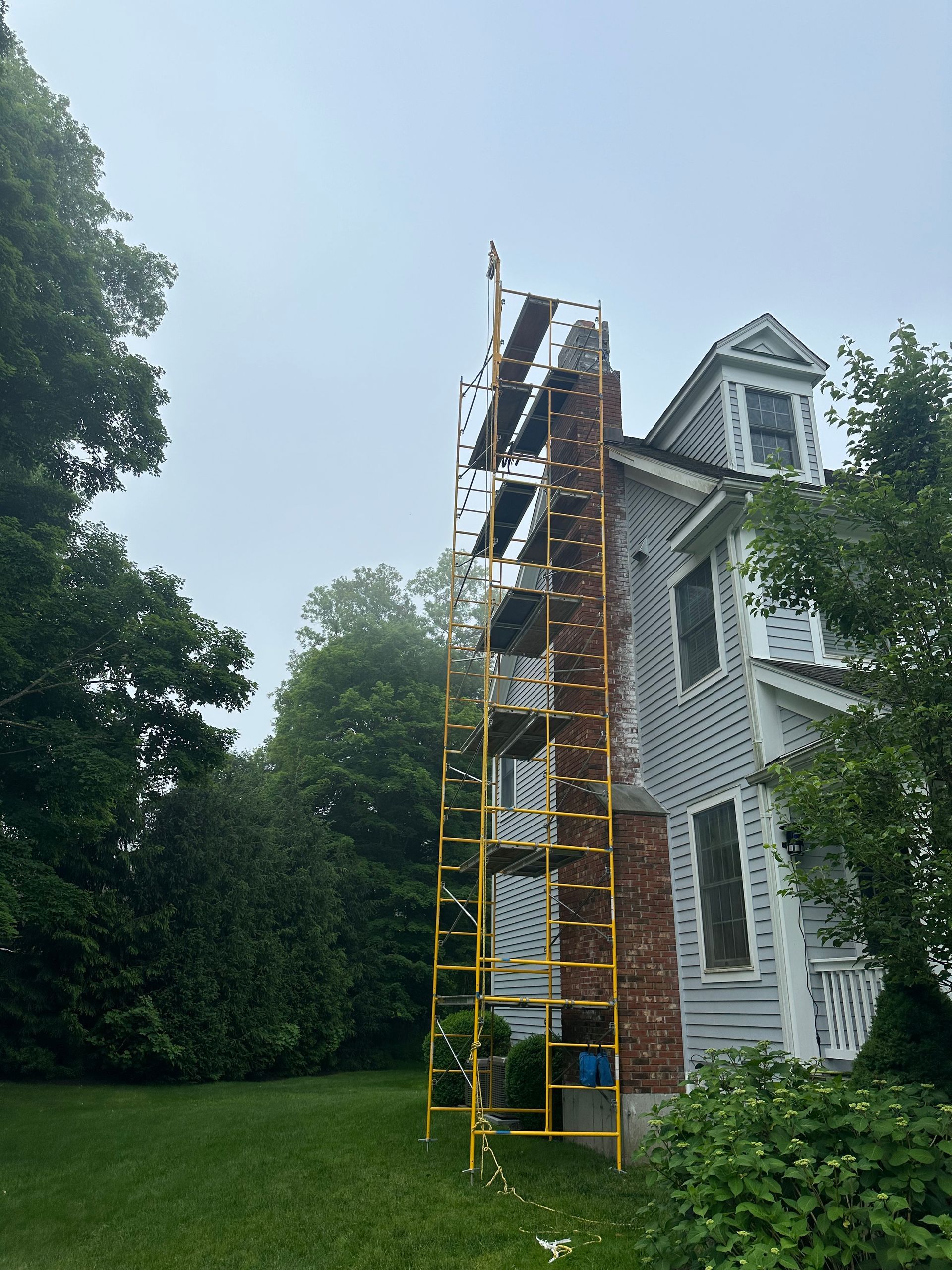 Yellow scaffolding stands beside a red brick chimney on the side of a white house surrounded by trees.
