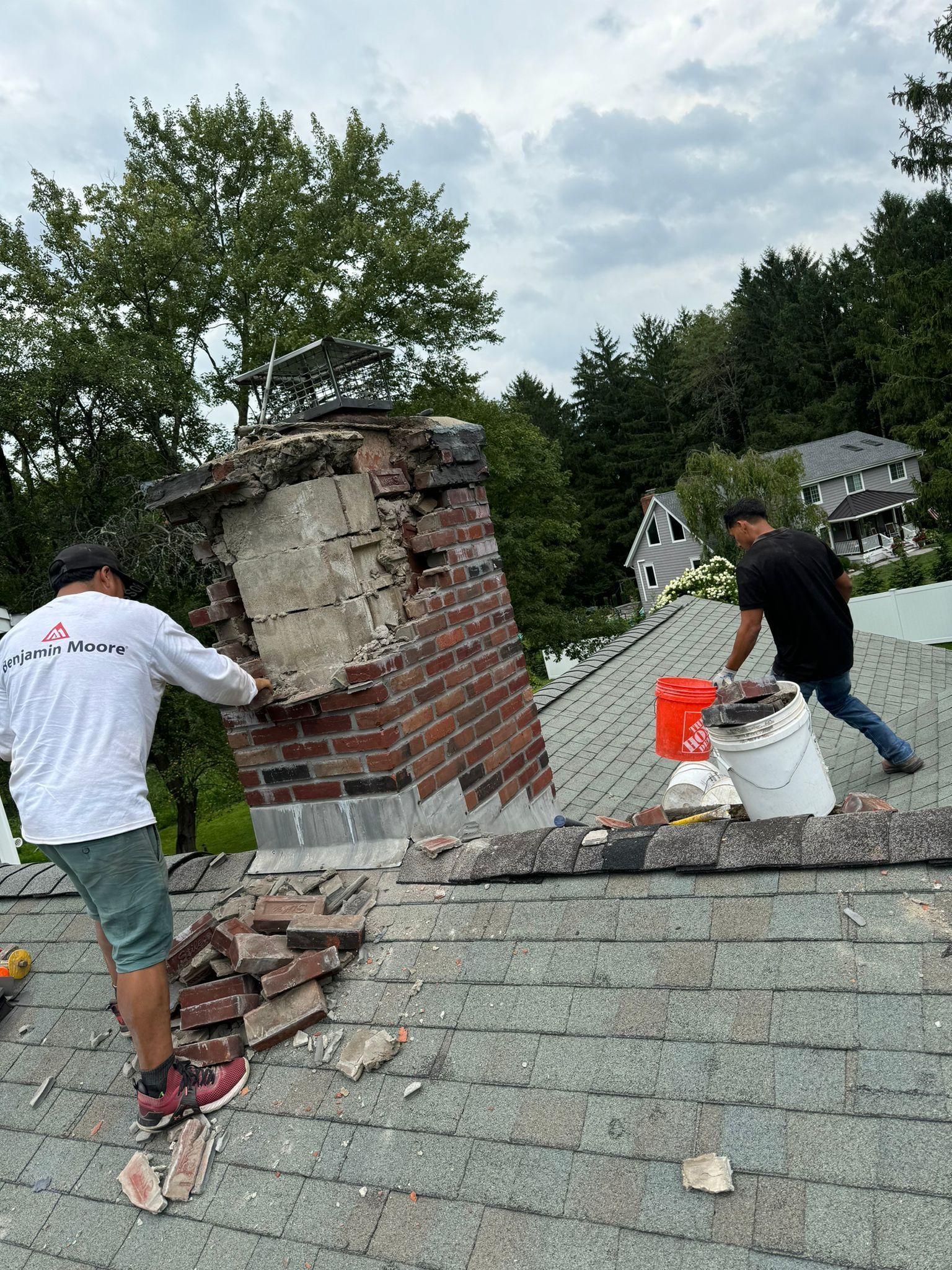 Two workers repair a damaged brick chimney on a shingled residential roof.