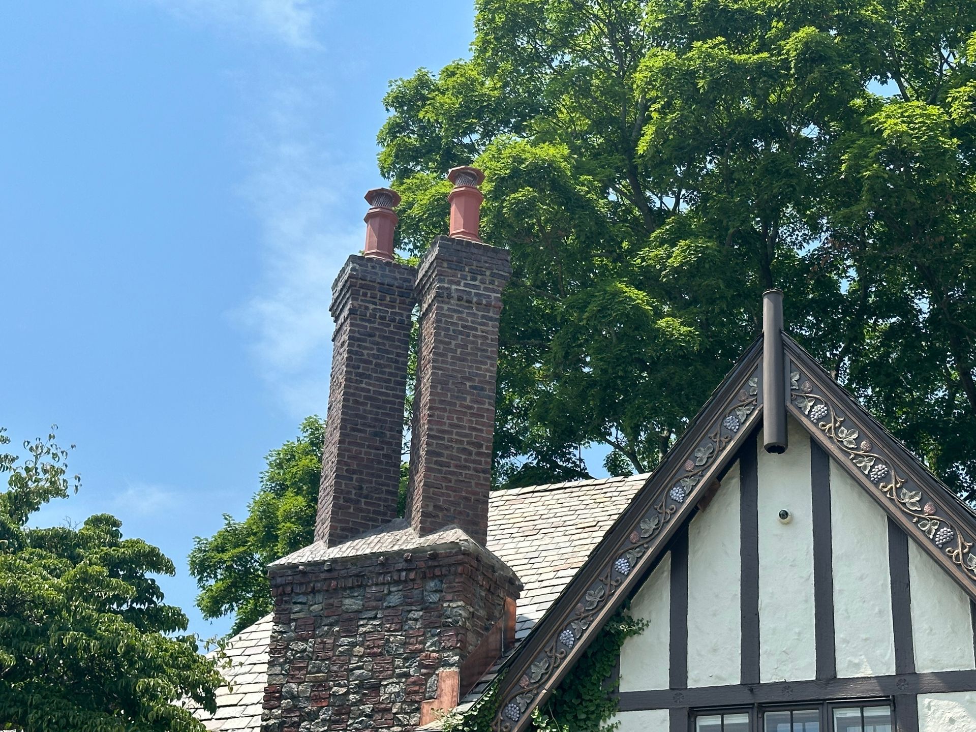 A brick chimney with two terracotta pots atop a Tudor-style home with white stucco walls, dark beams, and lush trees.