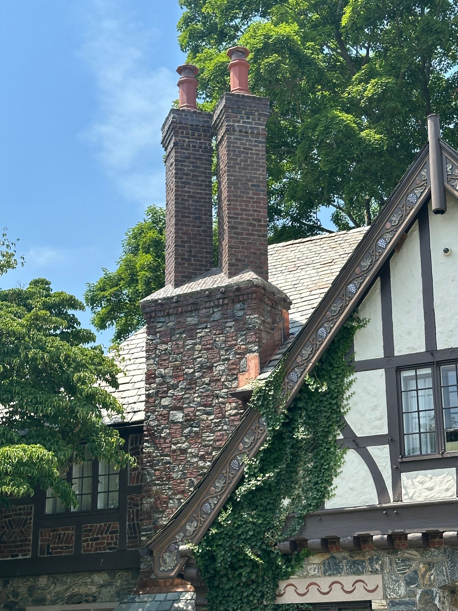 A fieldstone and brick chimney with two terracotta pots rises above a Tudor-style home with ivy and timber-framed walls.