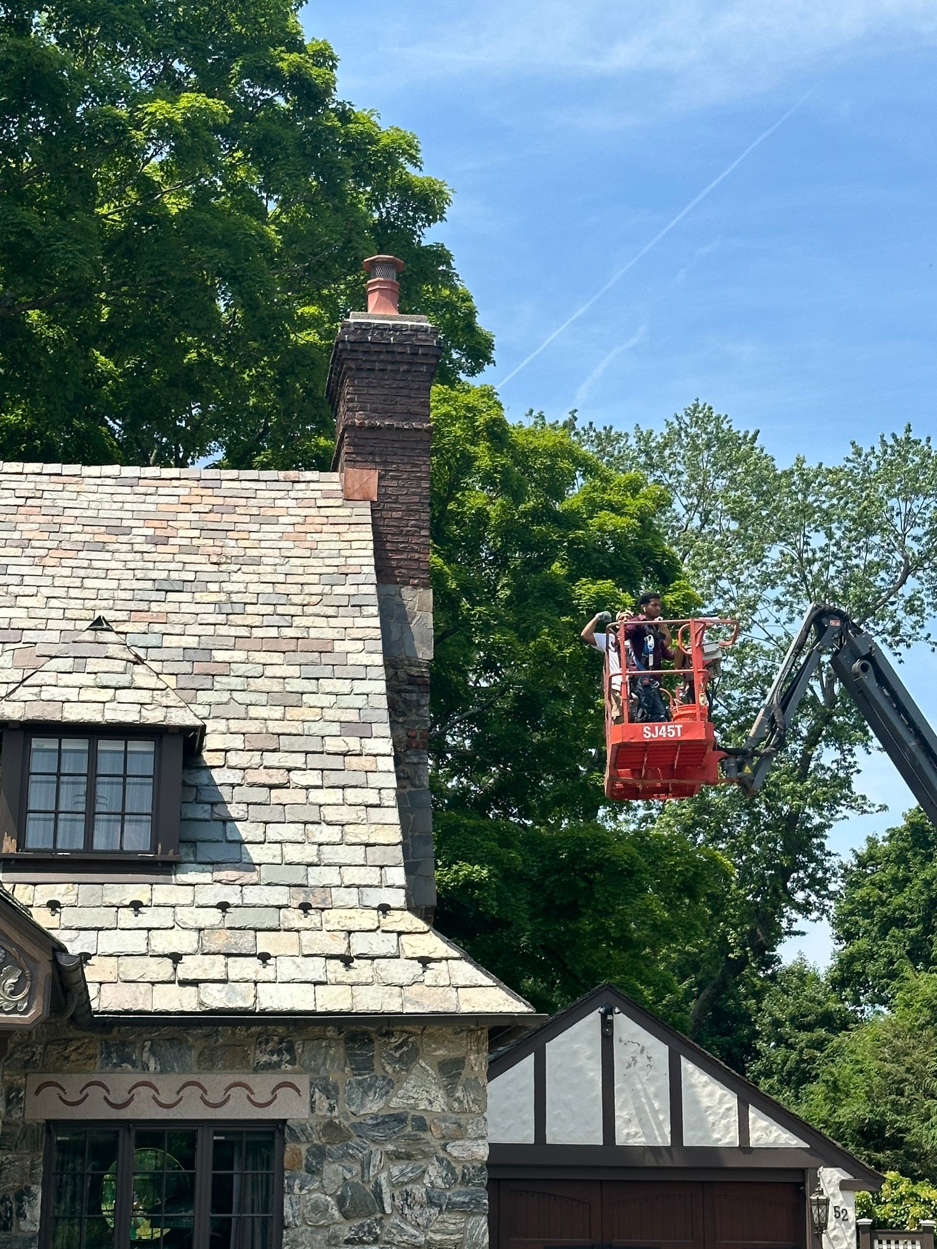 A worker in a red aerial lift platform works on the chimney of a stone house with a slate roof on a sunny day.