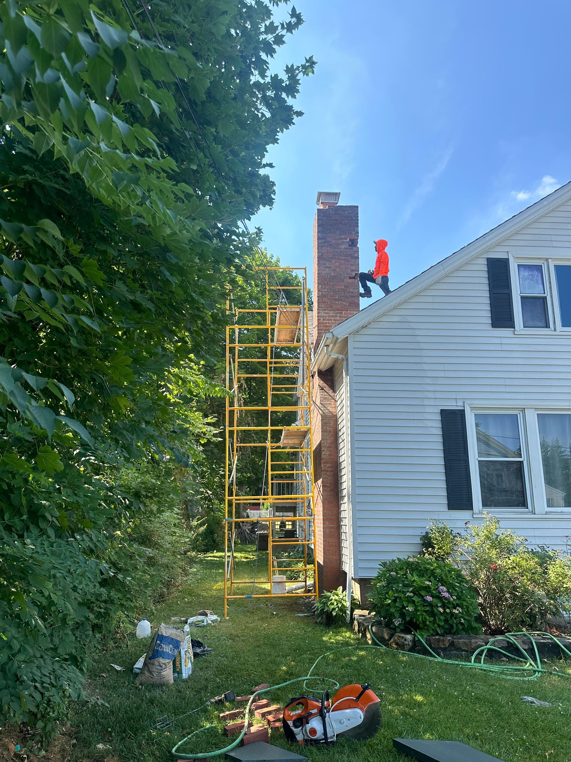 A person in a bright orange shirt works on a brick chimney from yellow scaffolding next to a white-sided house.