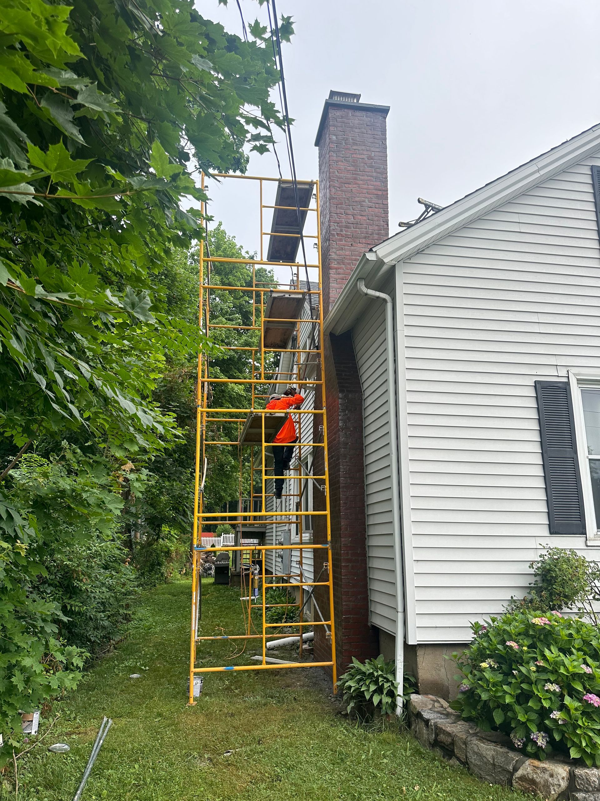 A person in an orange high-visibility jacket stands on yellow scaffolding to repair a brick chimney on a house.