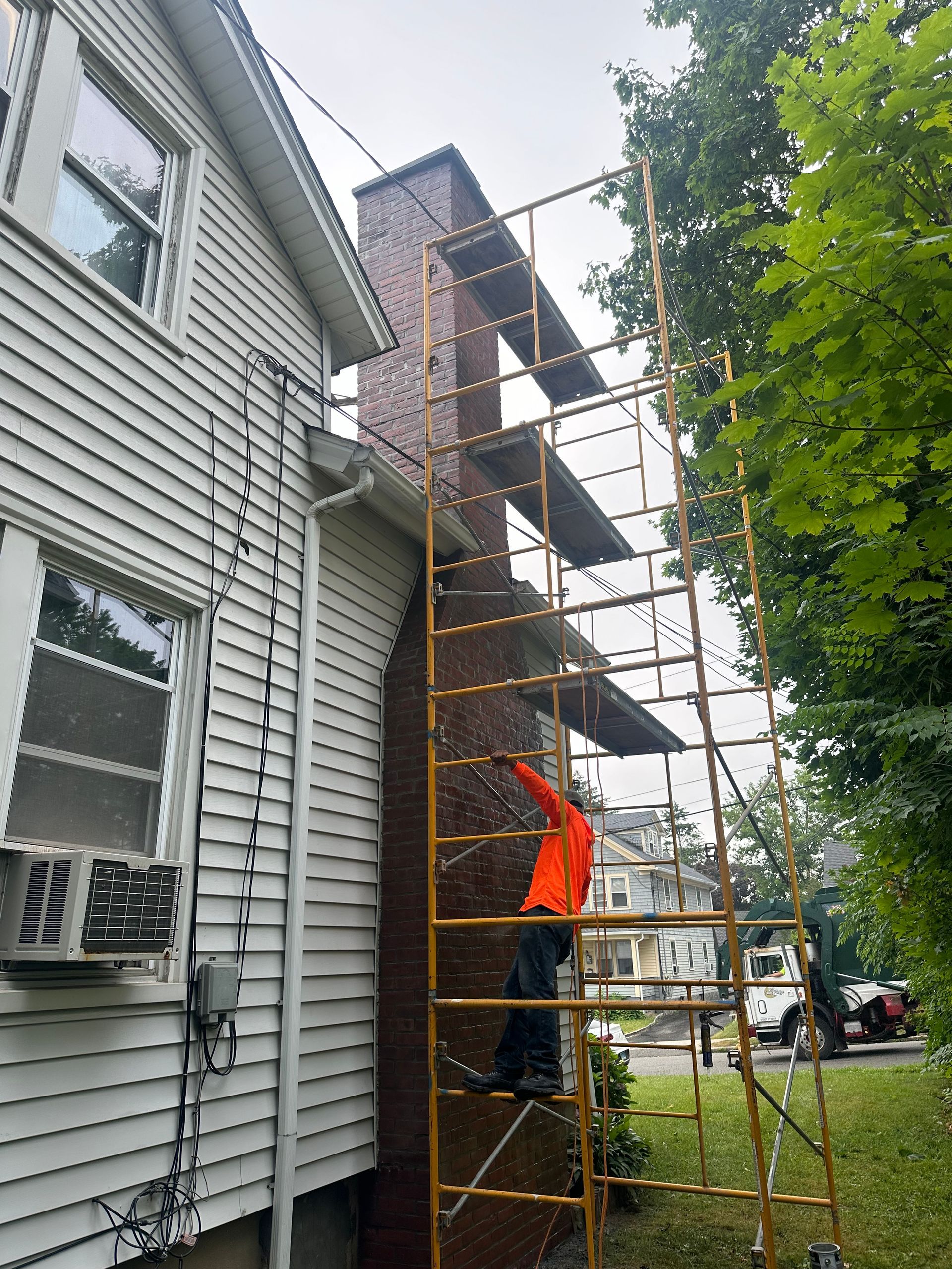 A construction worker in an orange shirt stands on metal scaffolding against a brick chimney on a house exterior.