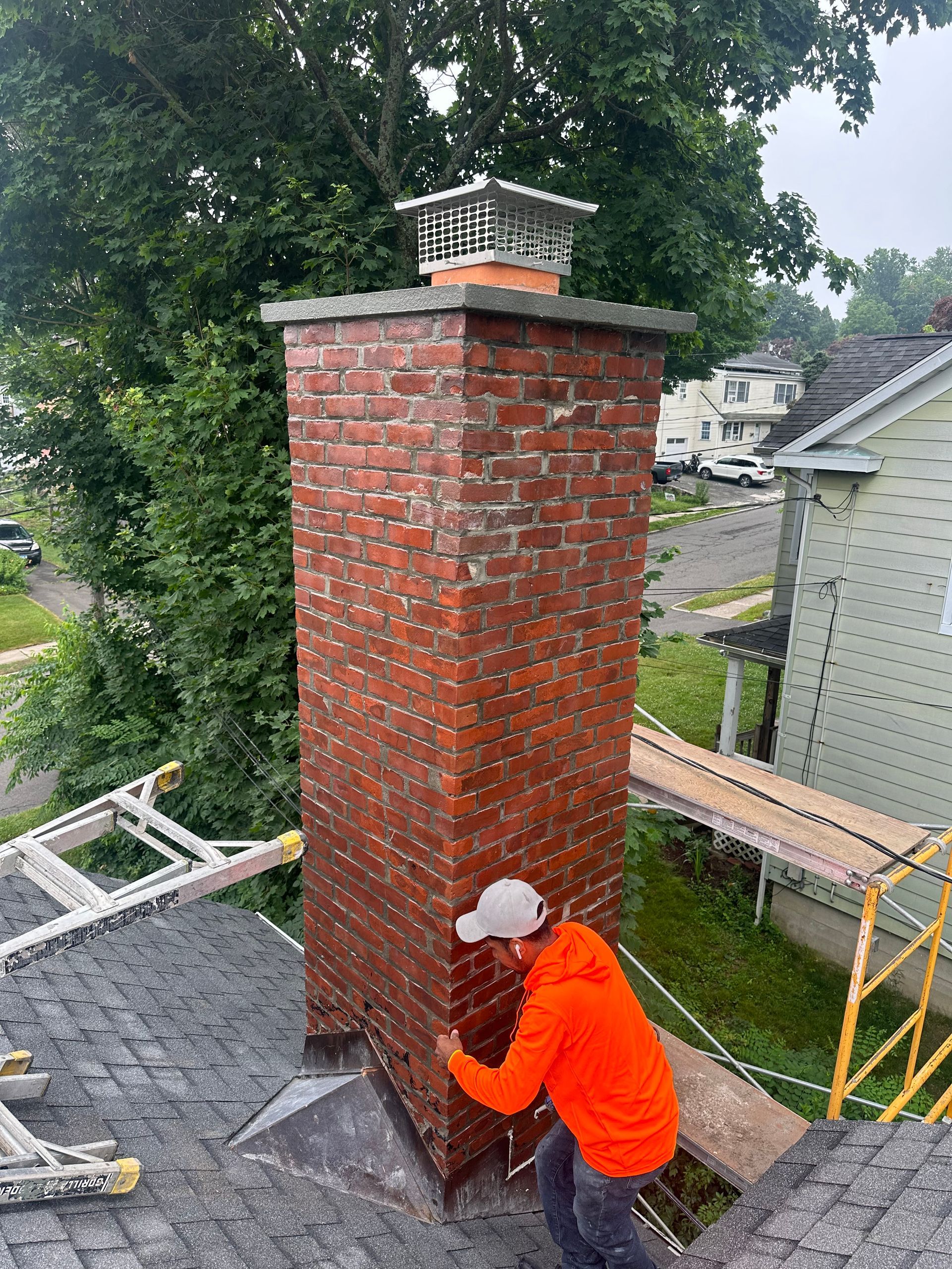 A person in an orange hoodie works on a brick chimney on a house roof.