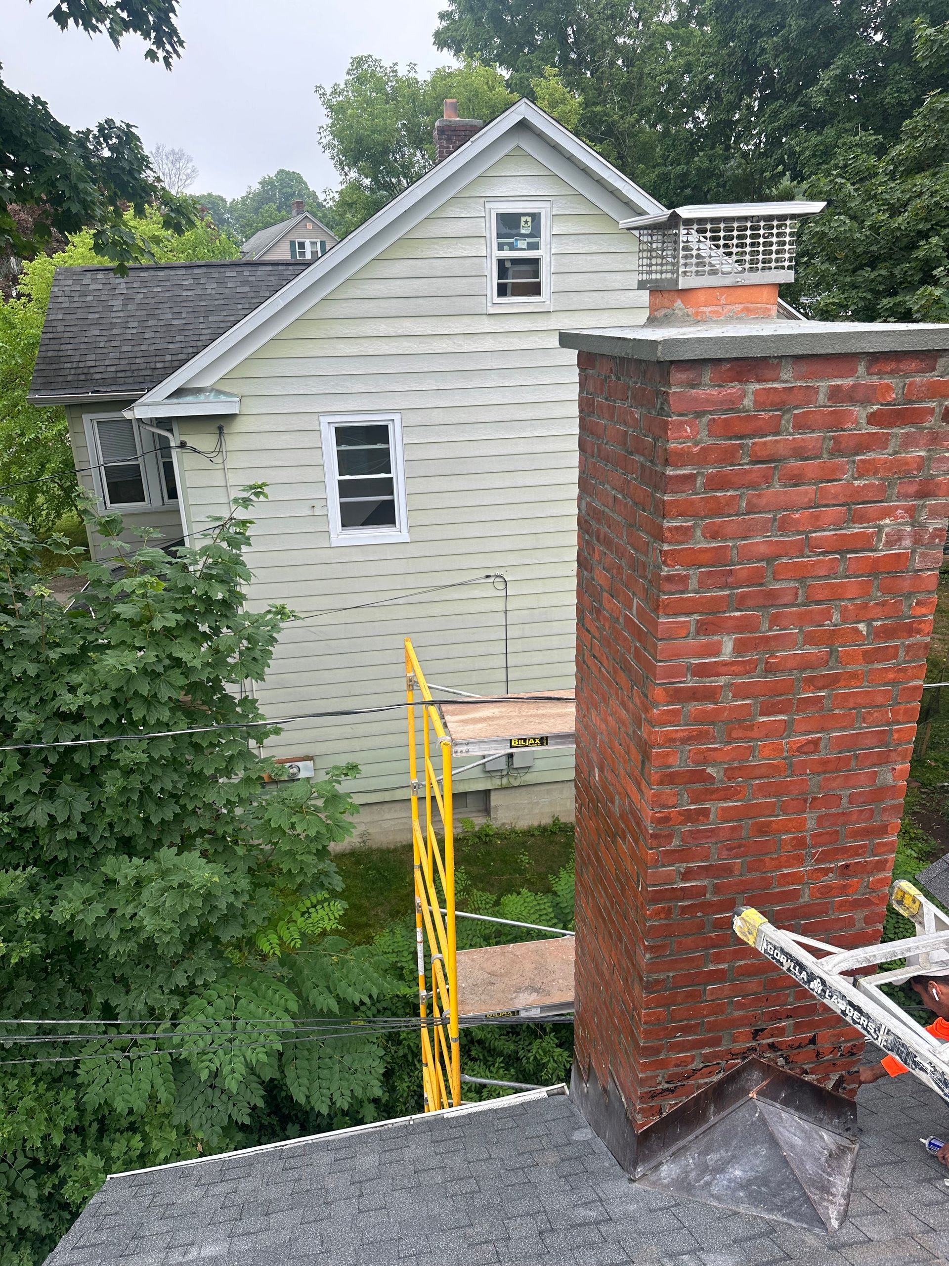 A high-angle view of a red brick chimney on a roof with scaffolding, overlooking a pale house with white trim.