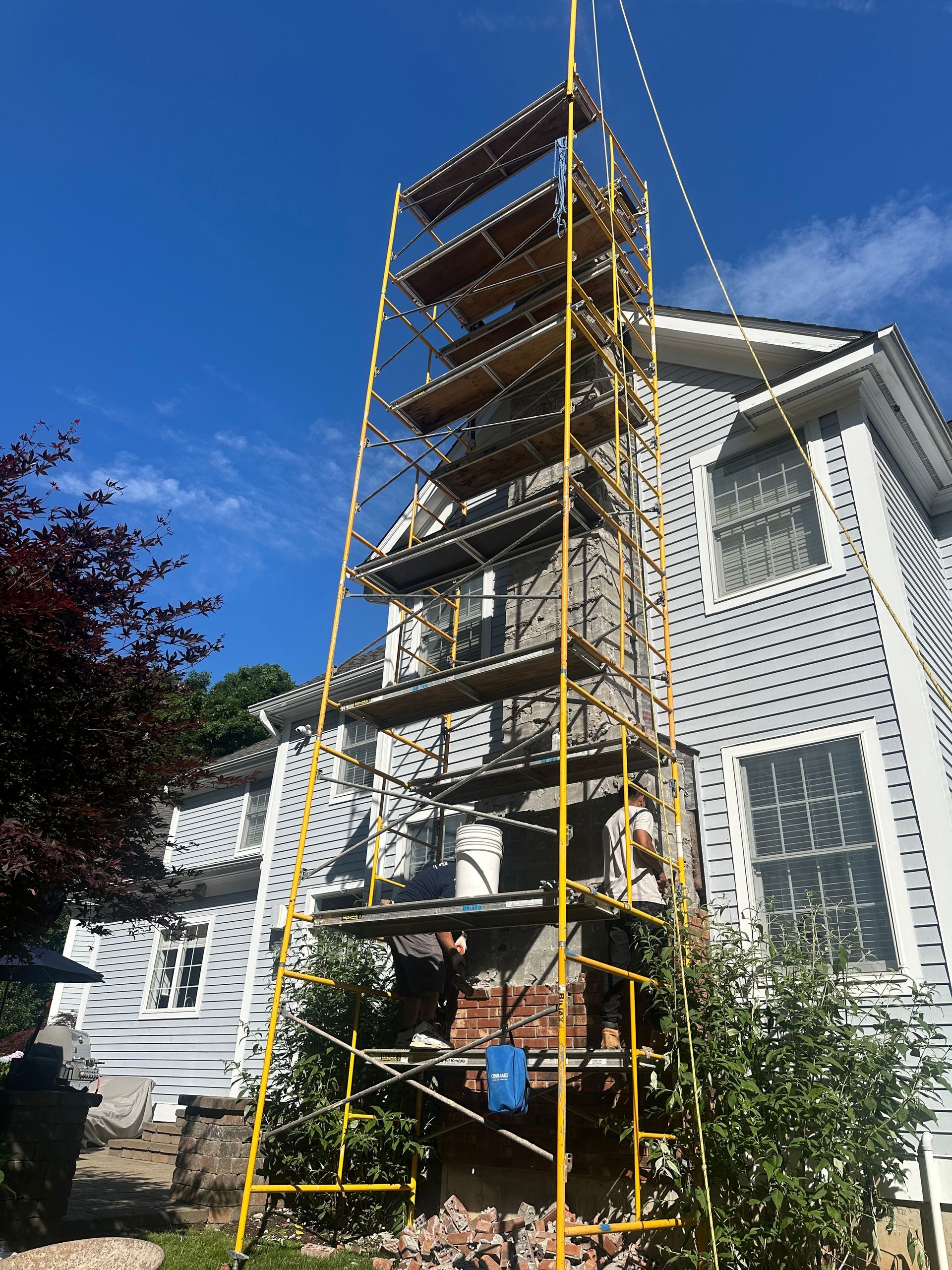 A tall yellow scaffolding tower stands against the side of a light-gray house, reaching toward the roof.