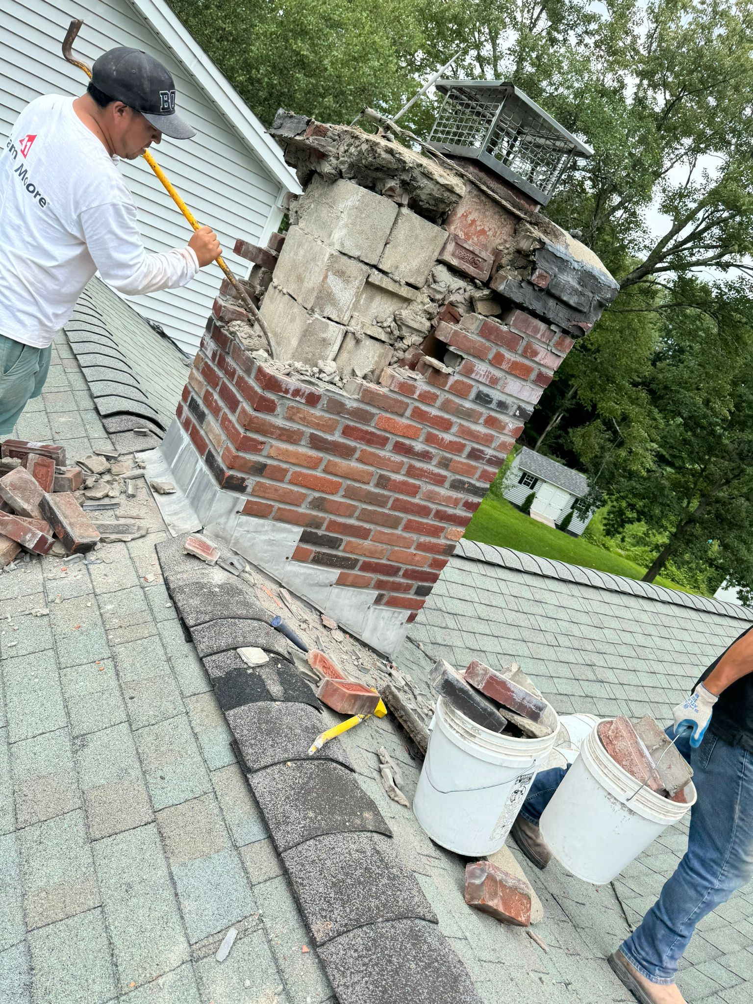 Two workers on a rooftop use tools and buckets to demolish a damaged brick chimney.