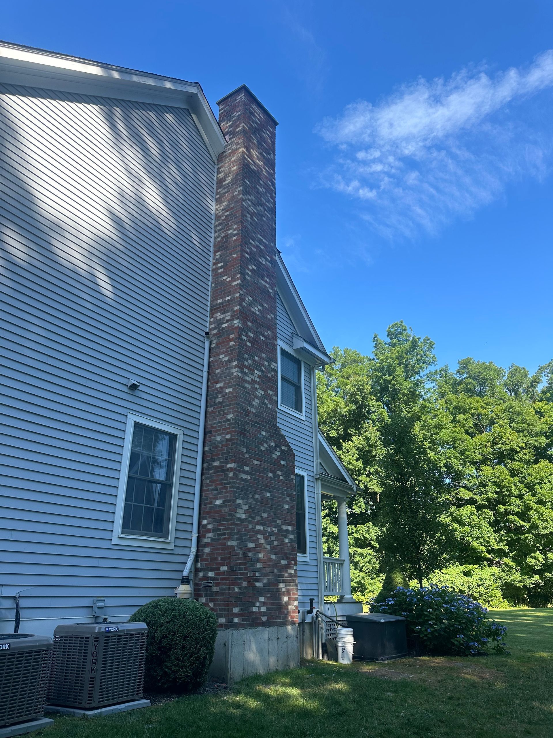 A multi-story house with light blue siding and a brick chimney standing against a clear blue sky and nearby trees.