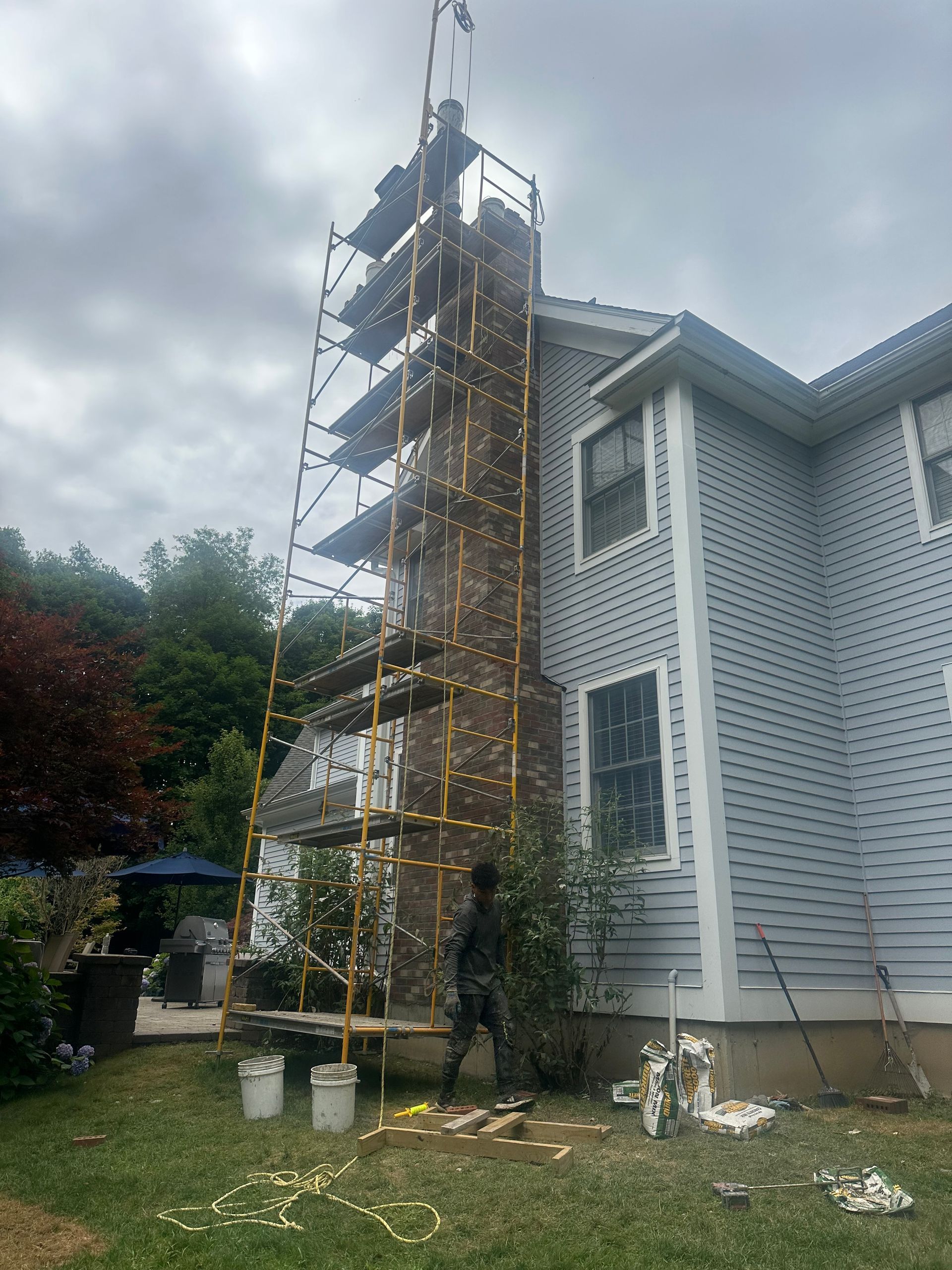 A person stands on the ground by a tall, multi-level scaffolding structure erected against a brick chimney on a house.