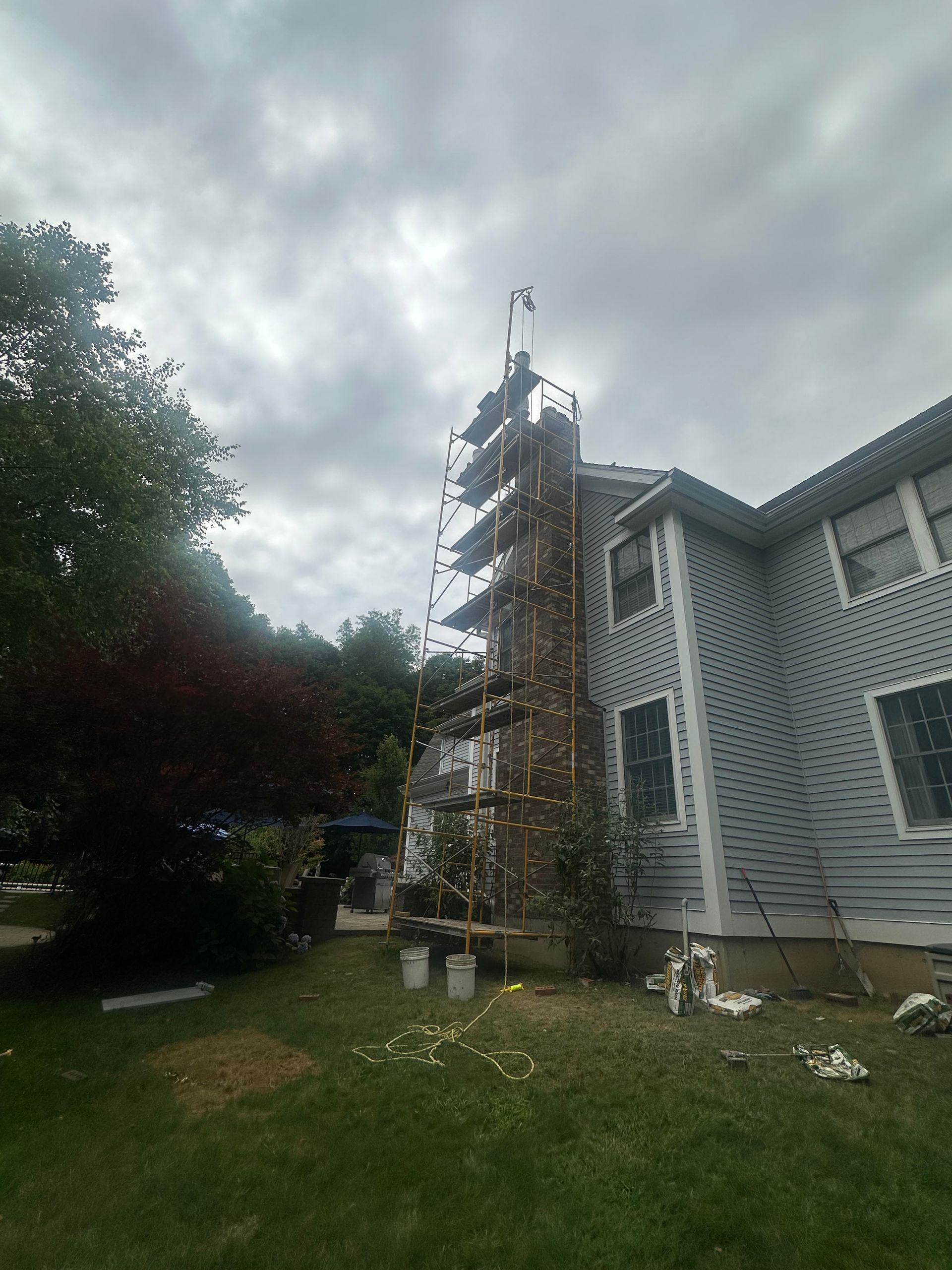 Scaffolding stands against a brick chimney on the side of a gray-sided house under a cloudy sky.