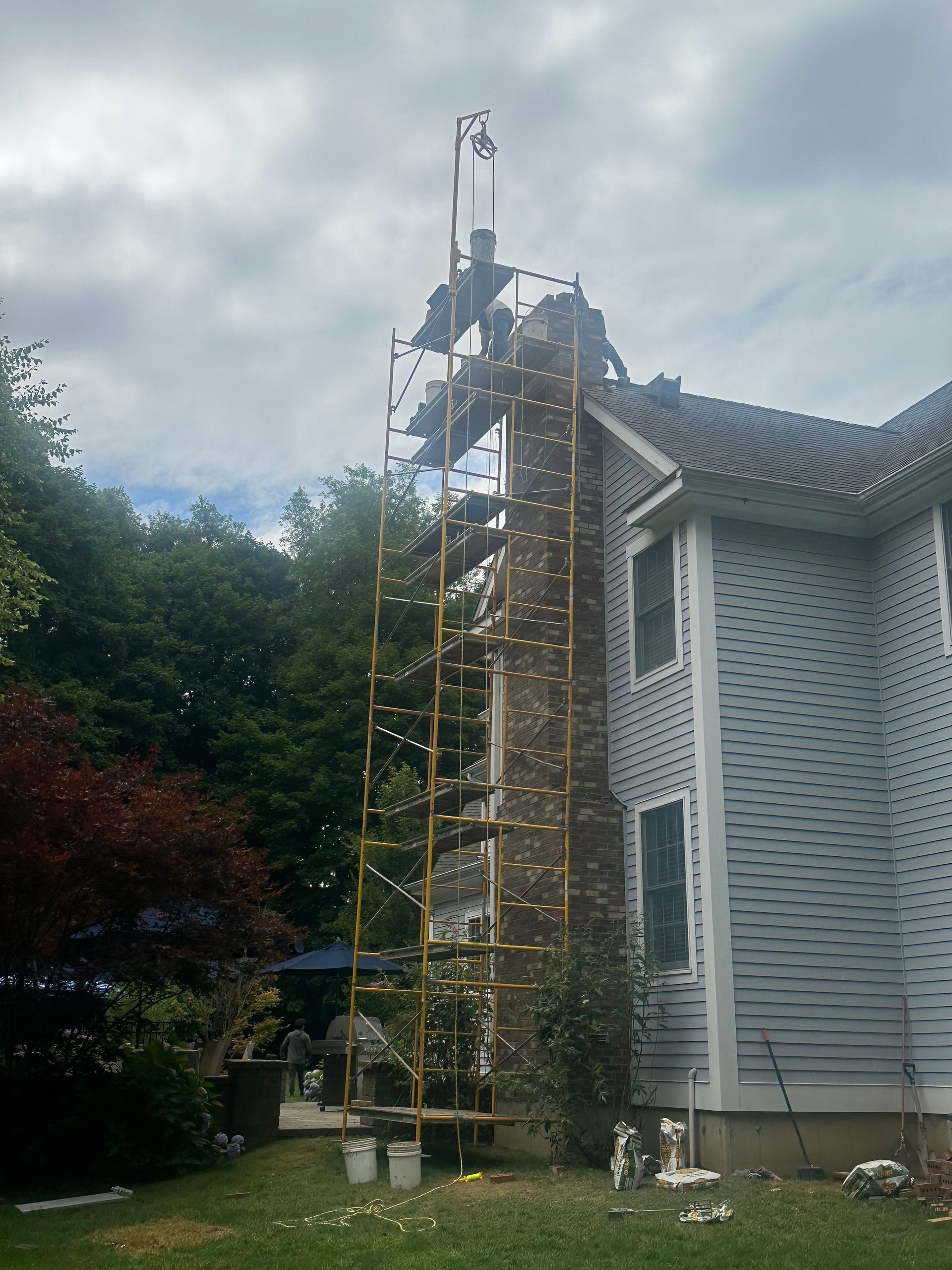 A tall yellow scaffold stands against a two-story house, reaching to the top of a brick chimney currently being repaired.