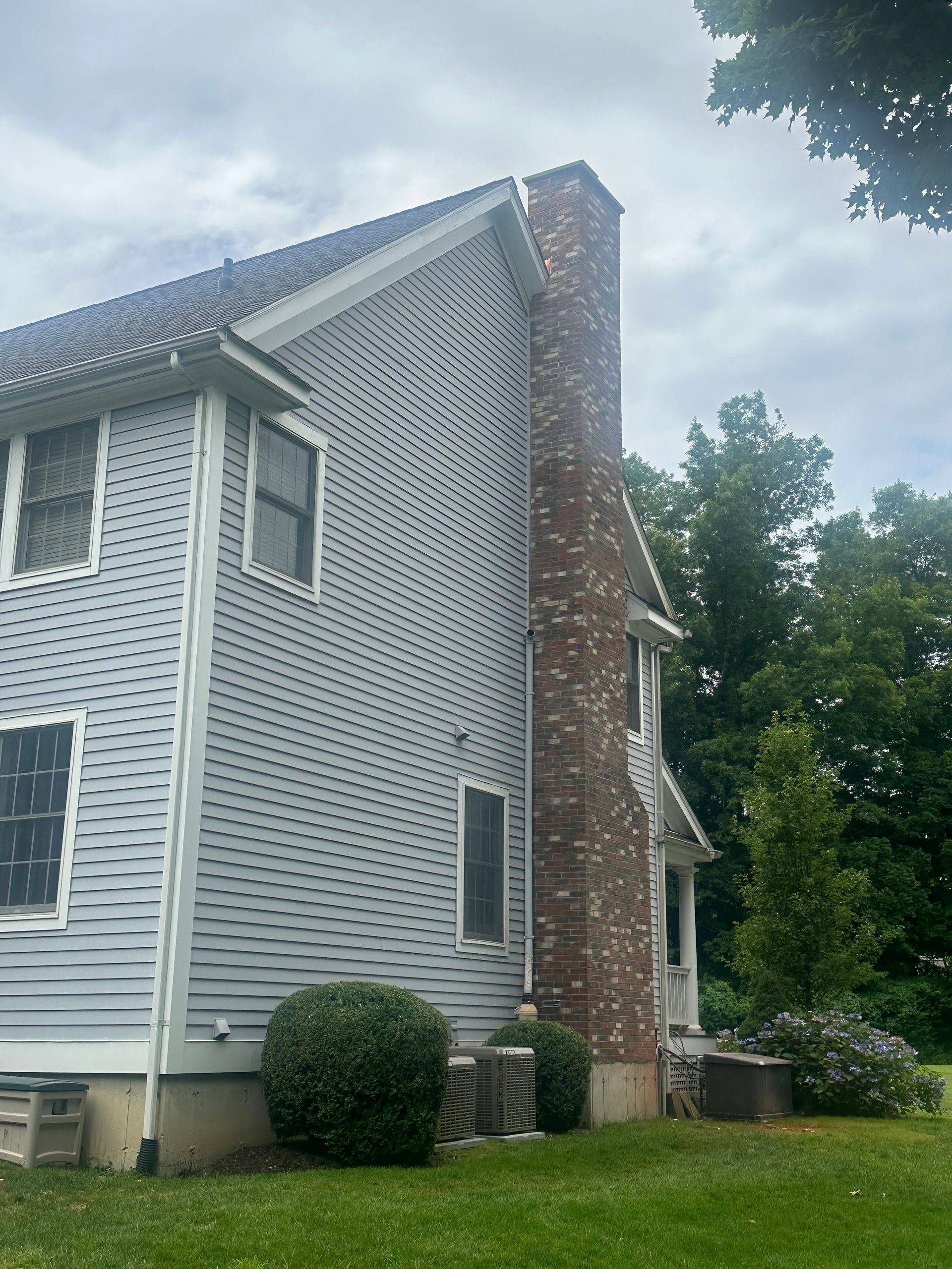 A side view of a two-story house with gray siding, a brick chimney, and surrounding greenery under a cloudy sky.